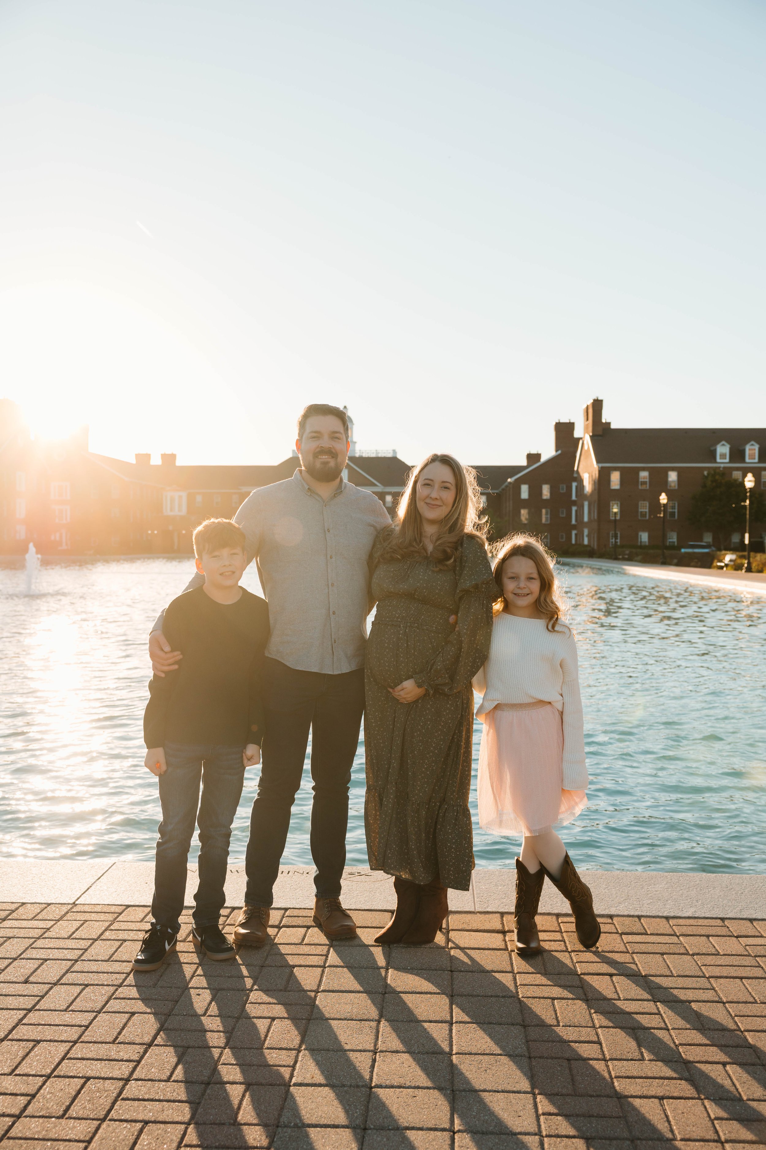 A family of four standing on a brick sidewalk by a water fountain with buildings in the background, bathed in warm sunlight.