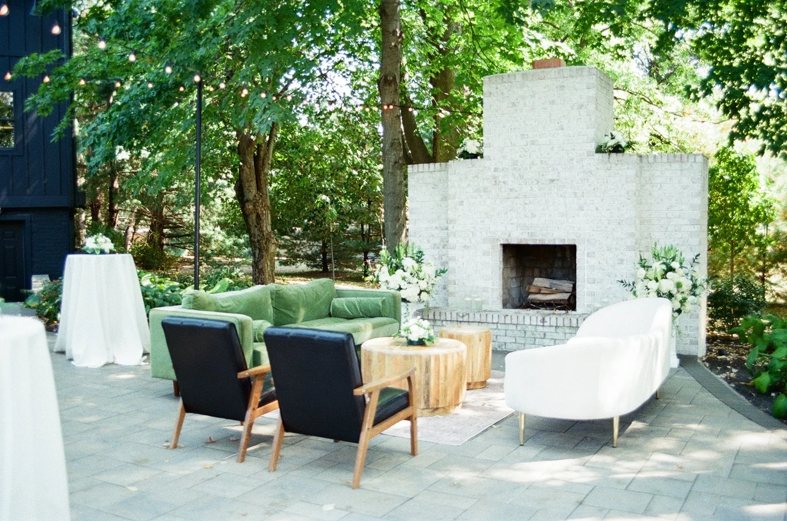 Outdoor patio with green sofa, white armchair, two black chairs, wooden coffee table, white fireplace, and string lights in a garden setting with trees and plants.