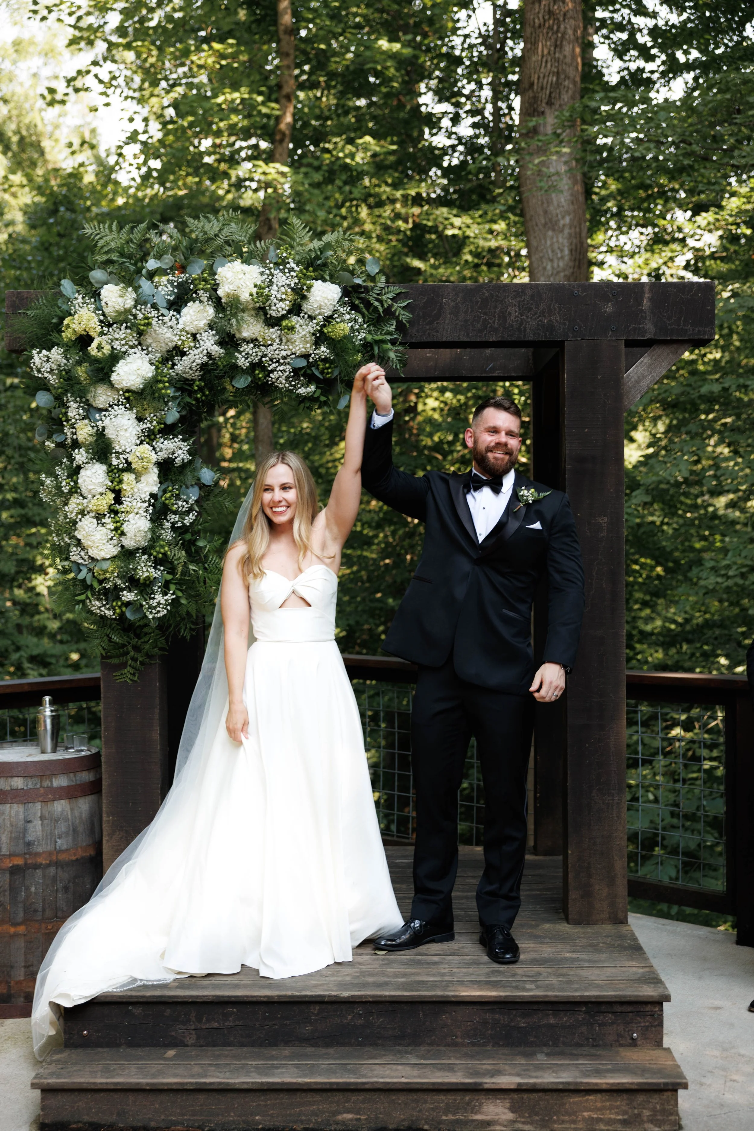 Bride and groom at their wedding, holding hands and smiling under a wooden arch decorated with white flowers and greenery, in an outdoor setting surrounded by trees.