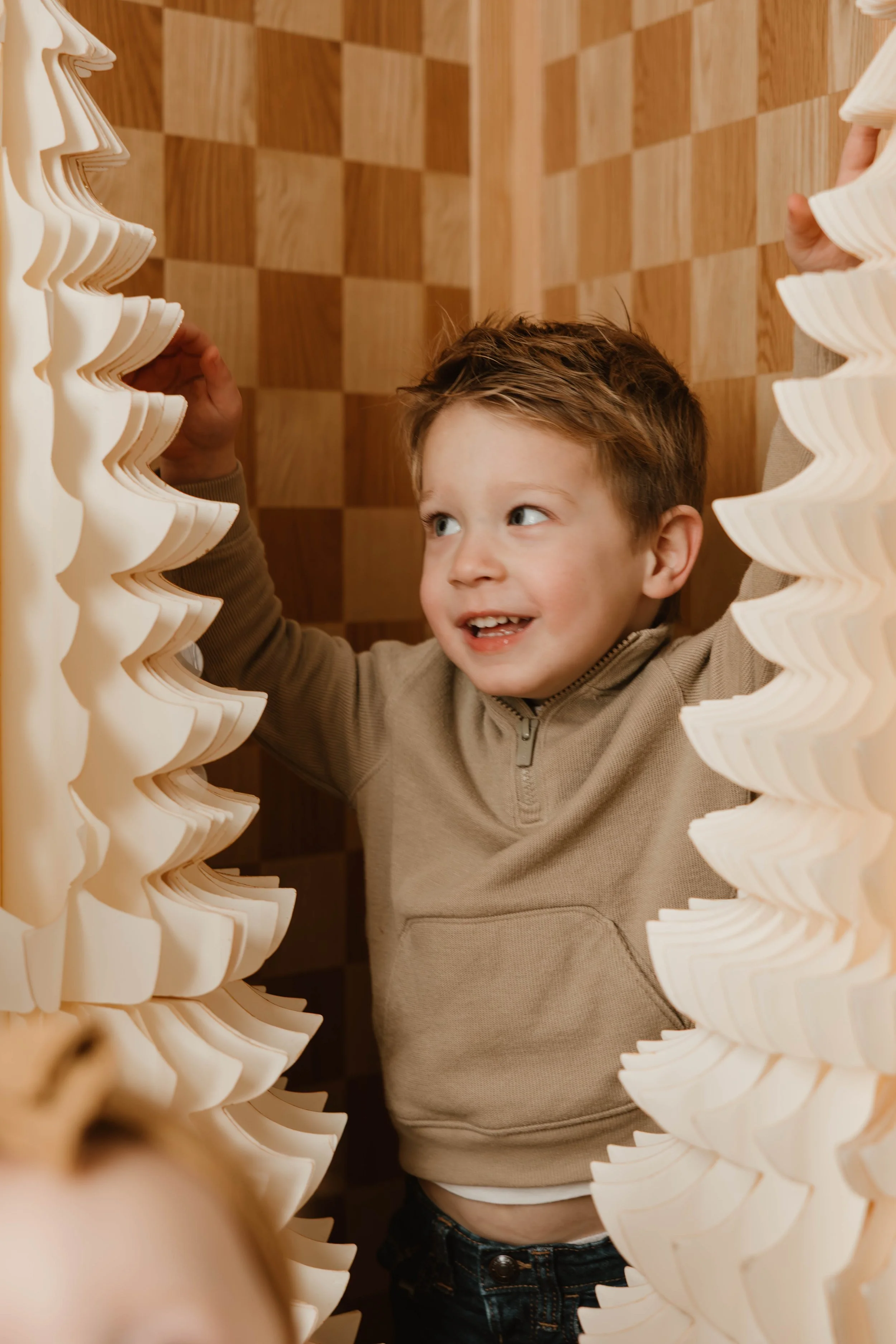 Smiling young boy with short, tousled hair, wearing a beige zip-up sweater, standing between two tall stacks of white foam or plastic trays on a checkered wooden wall background.
