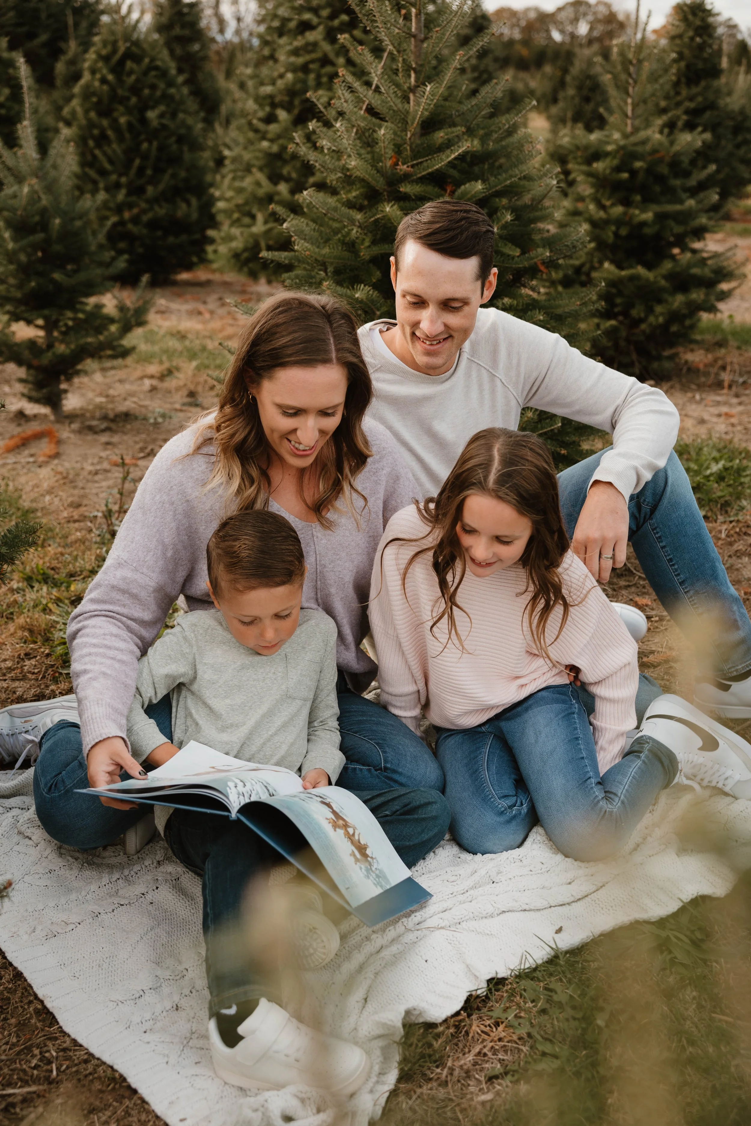 Family of four, two adults and two children, sitting on a blanket outdoors in a Christmas tree farm, looking at a book together.