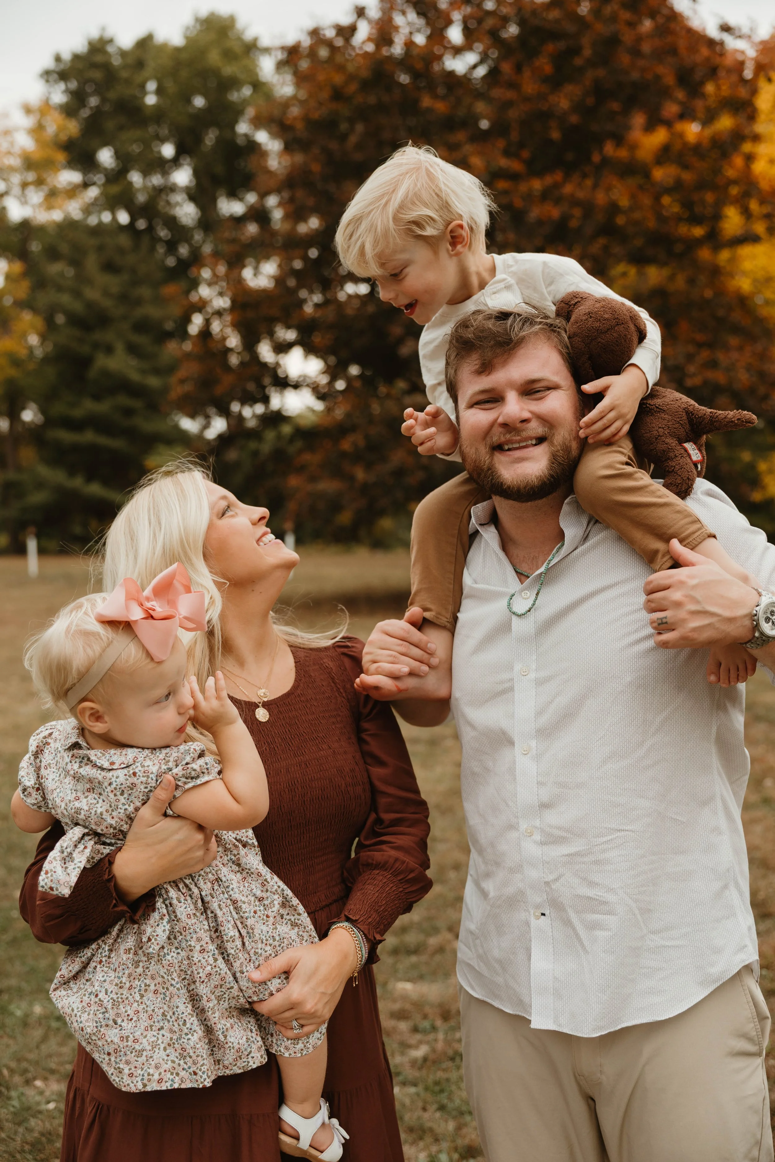 A family of four outdoors in autumn, with colorful fall trees in the background. The father is carrying his young son on his shoulders, and the mother is holding their toddler daughter.