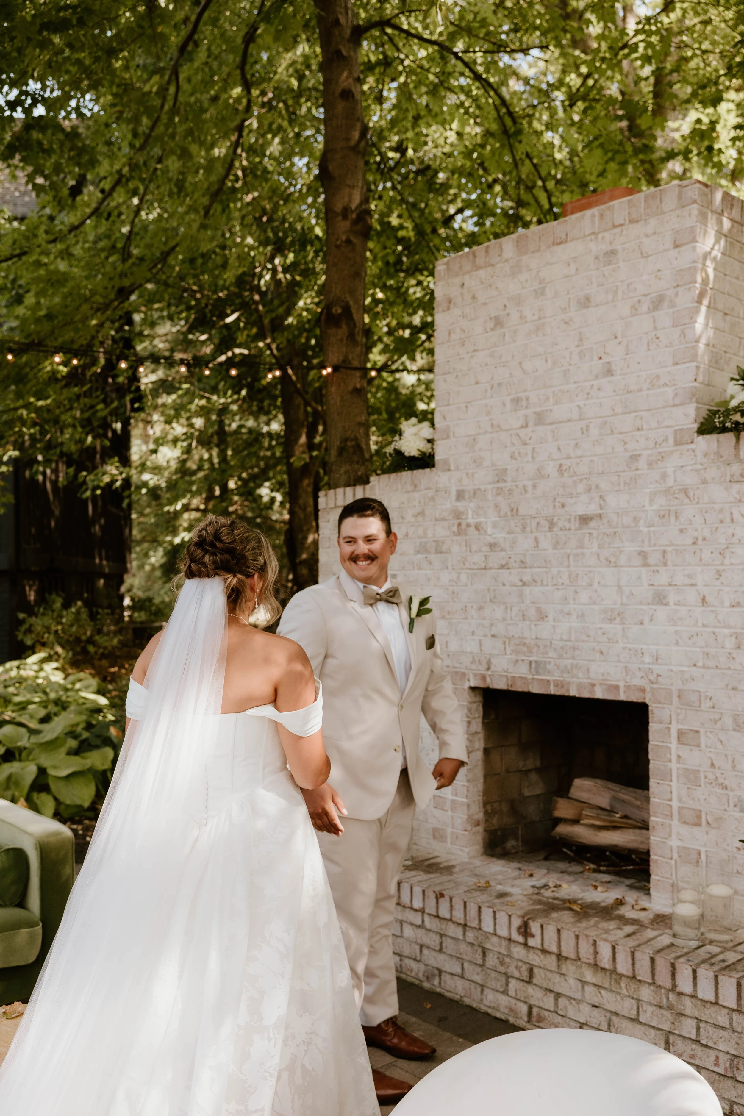 A bride and groom smiling at each other during their wedding ceremony outdoors, with green trees and string lights in the background.