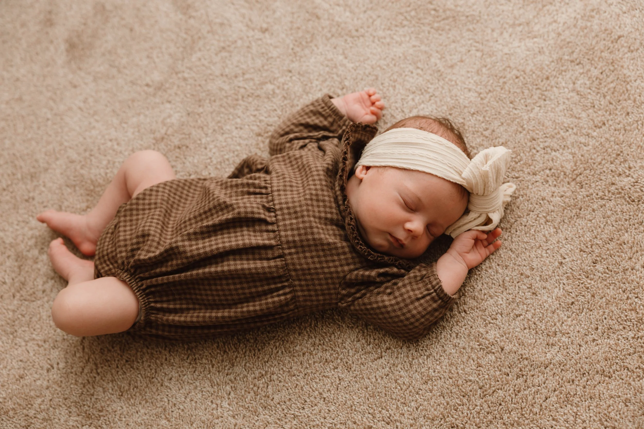 A sleeping baby girl lying on a beige carpet, wearing a brown checkered dress and a cream-colored headband with a bow.