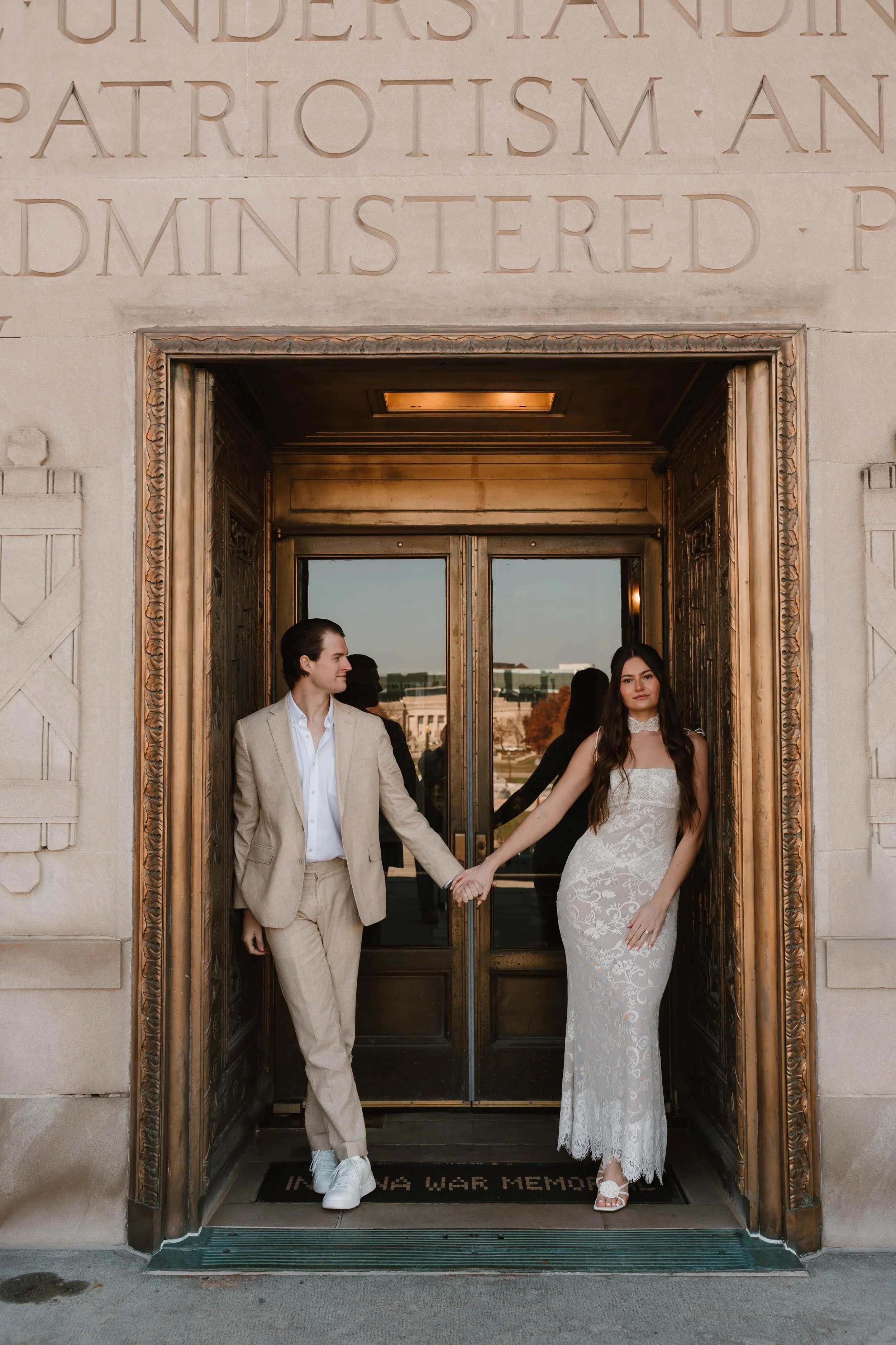 A man and woman holding hands in front of a building entrance with glass doors. The man is dressed in a beige suit, and the woman is wearing a long white lace dress. They are standing on a doormat that reads "IN LOVING MEMORY OF".