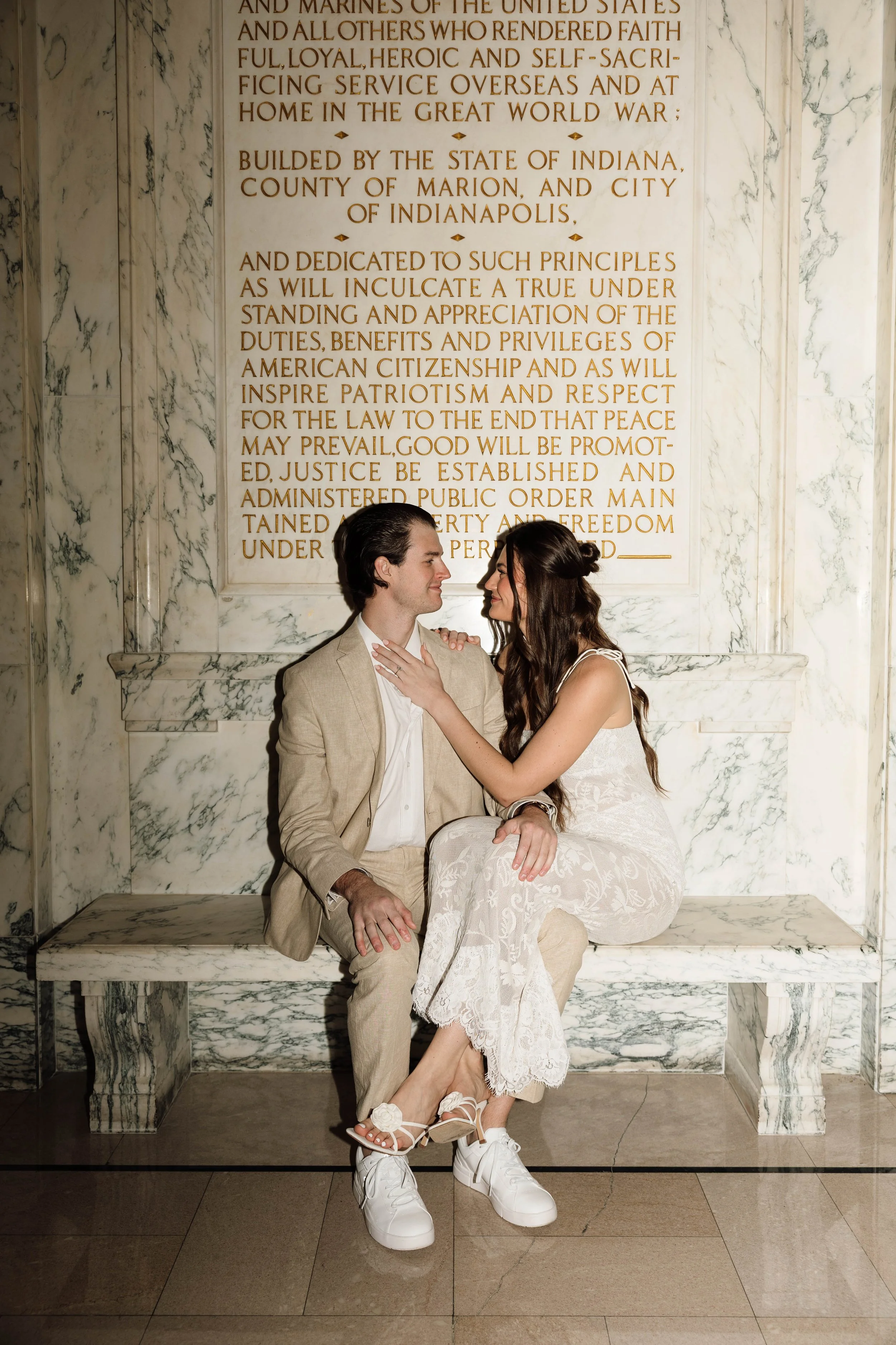 A couple sitting on a marble bench in front of a memorial with a large engraved text plaque, inside a marble hall.