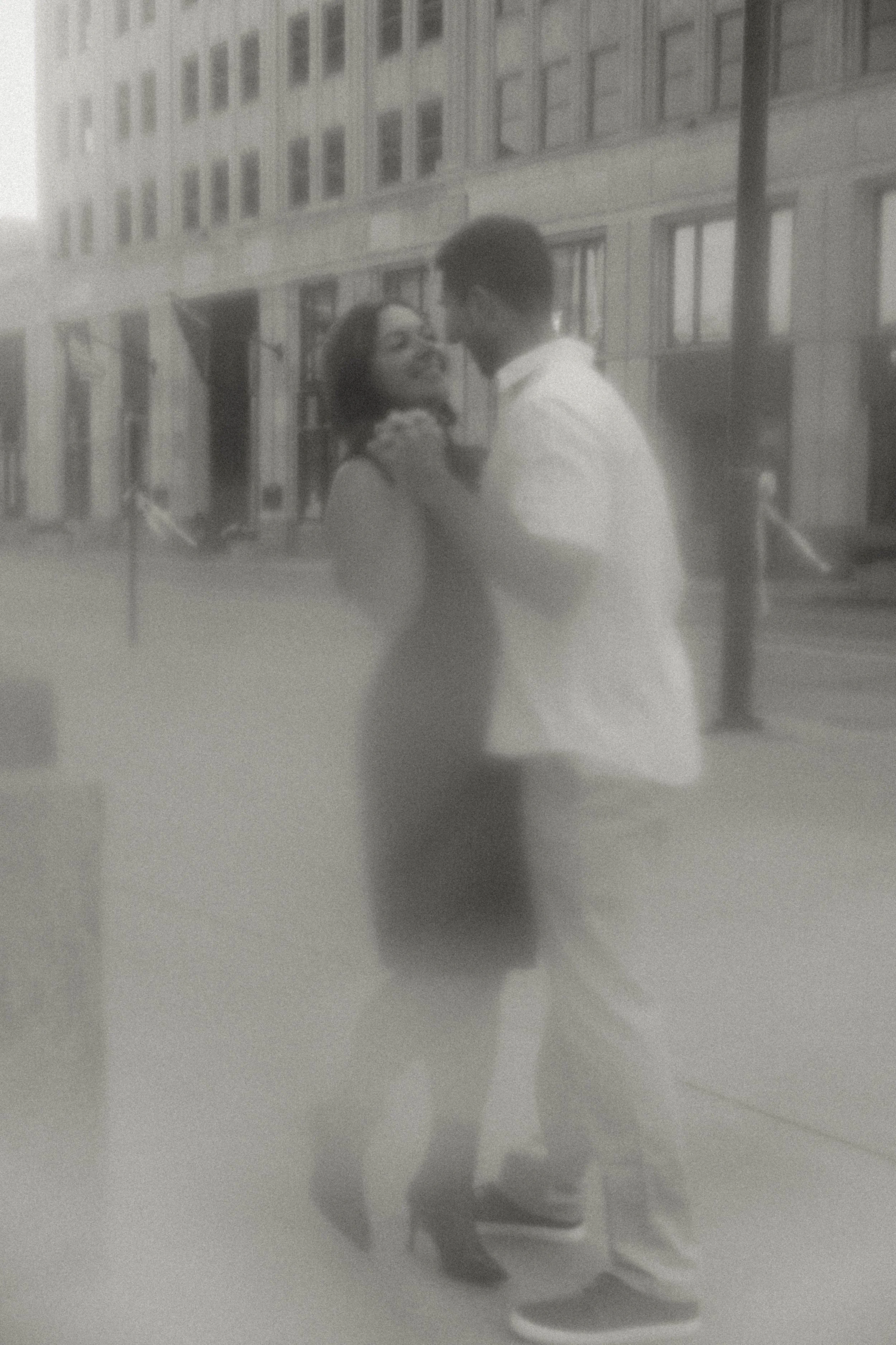 A black and white photo of a couple dancing outdoors on a city street, with buildings in the background.