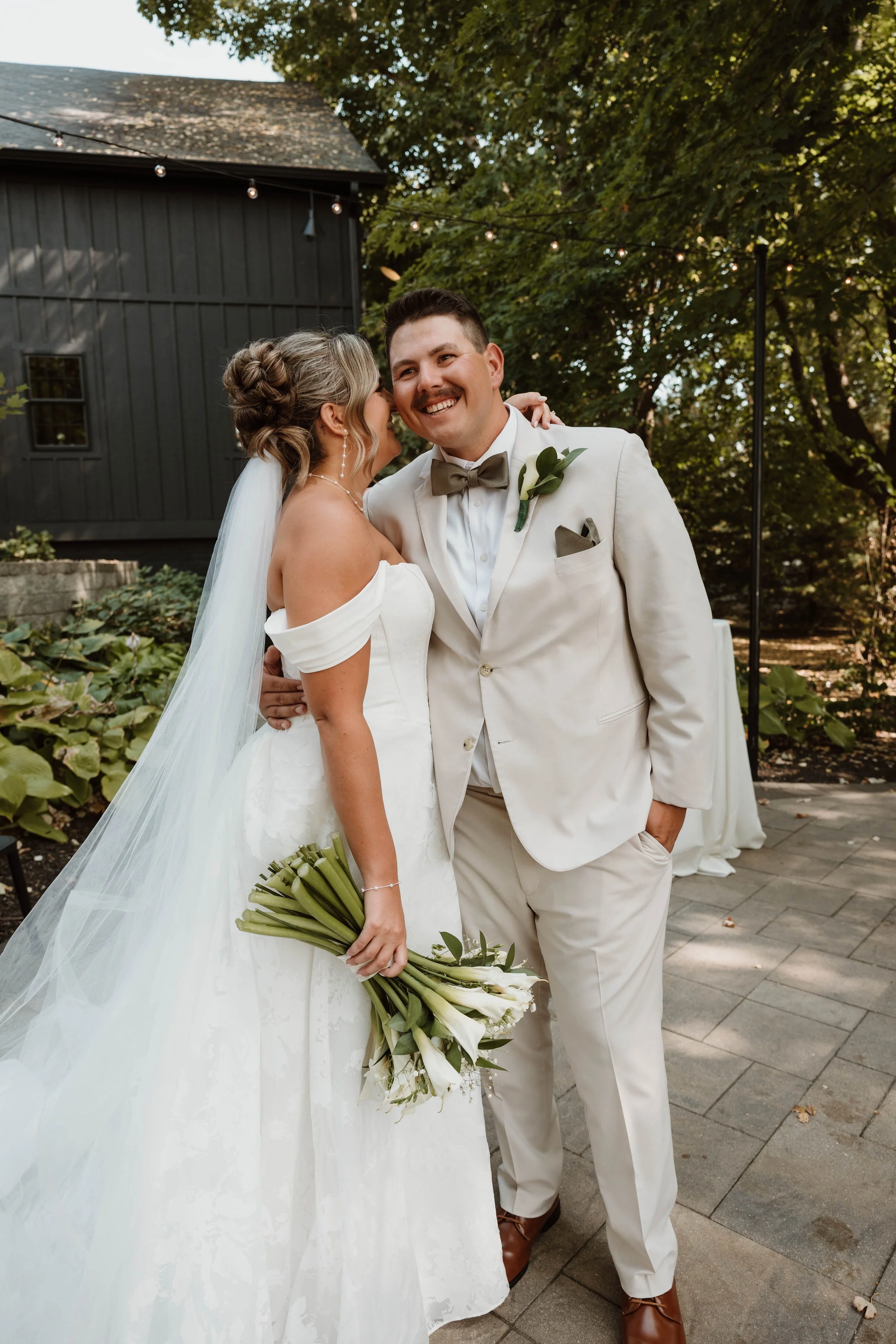 A bride and groom at their wedding, standing outdoors, smiling and embracing each other. The bride holds a bouquet of white flowers, and the groom is dressed in a cream suit with a bow tie and boutonniere. The setting includes trees and string lights