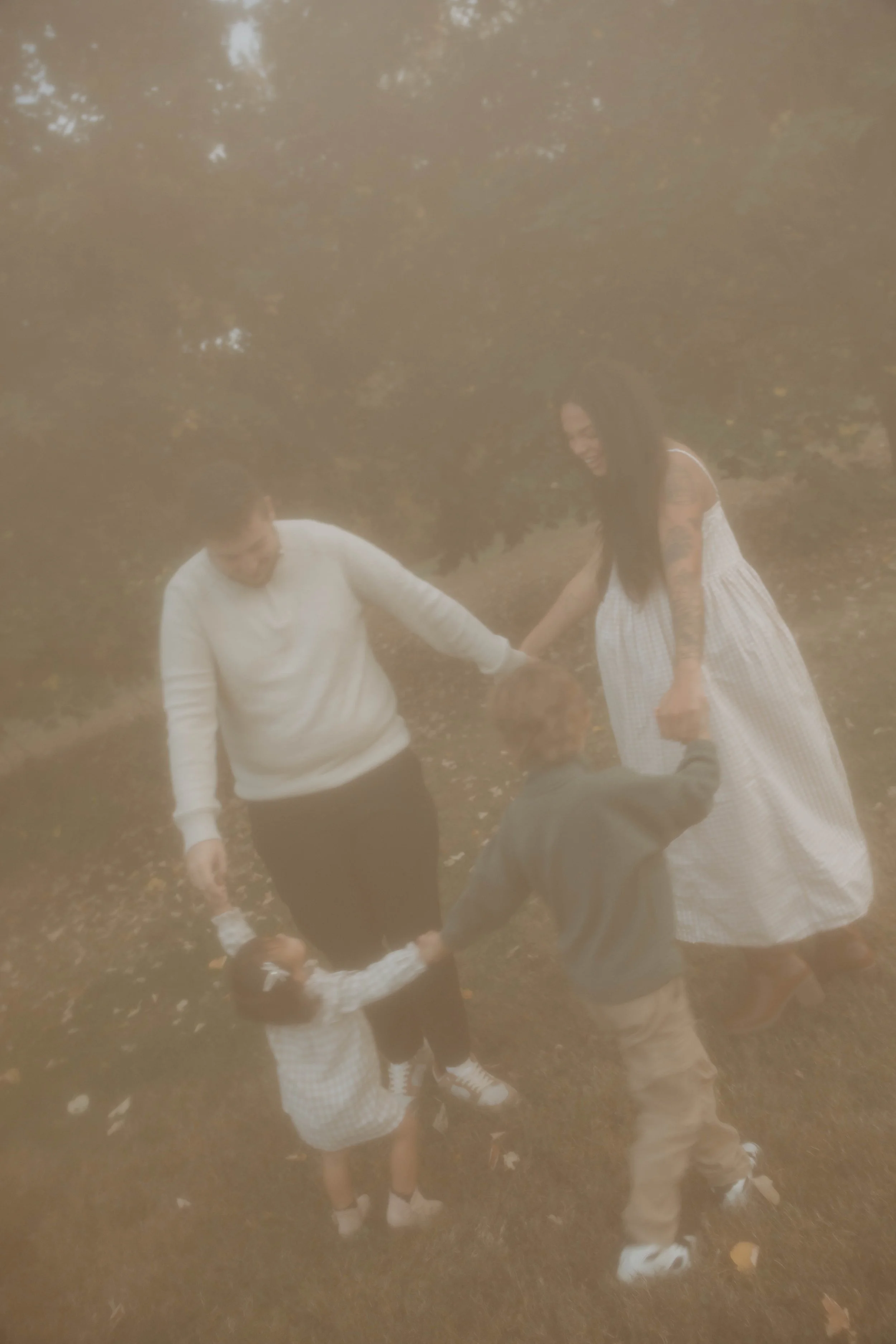 A hazy, foggy outdoor scene where four people, including two children and two adults, are holding hands in a circle, smiling and playing.