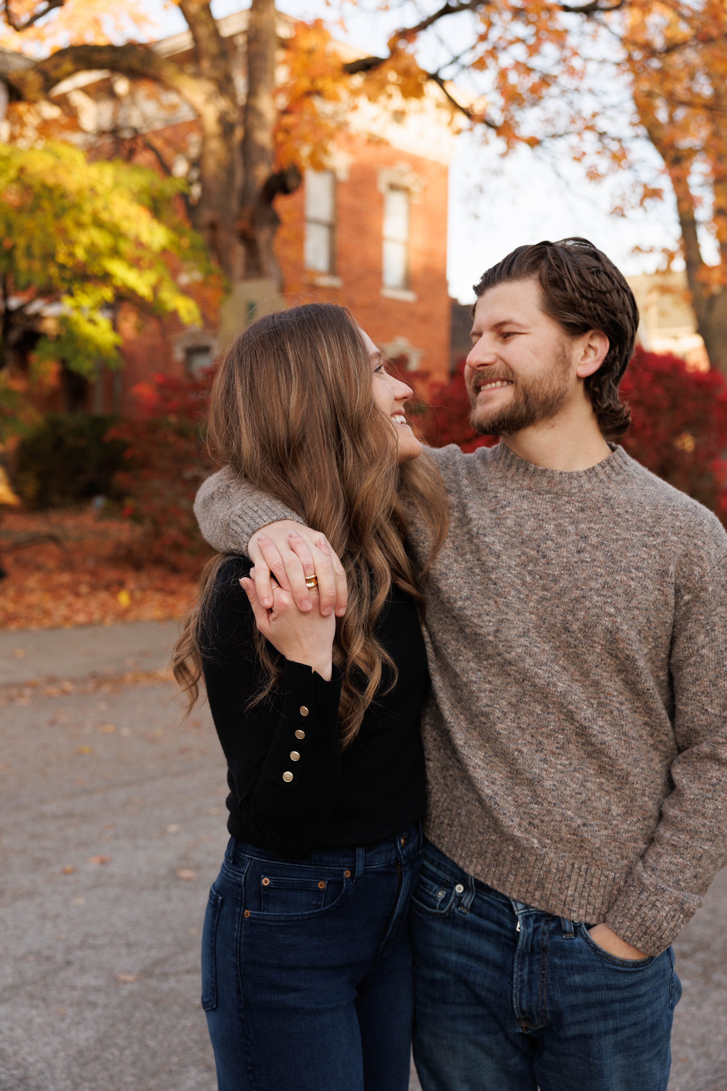 A smiling couple standing close together outdoors during autumn, with trees and a brick house in the background.