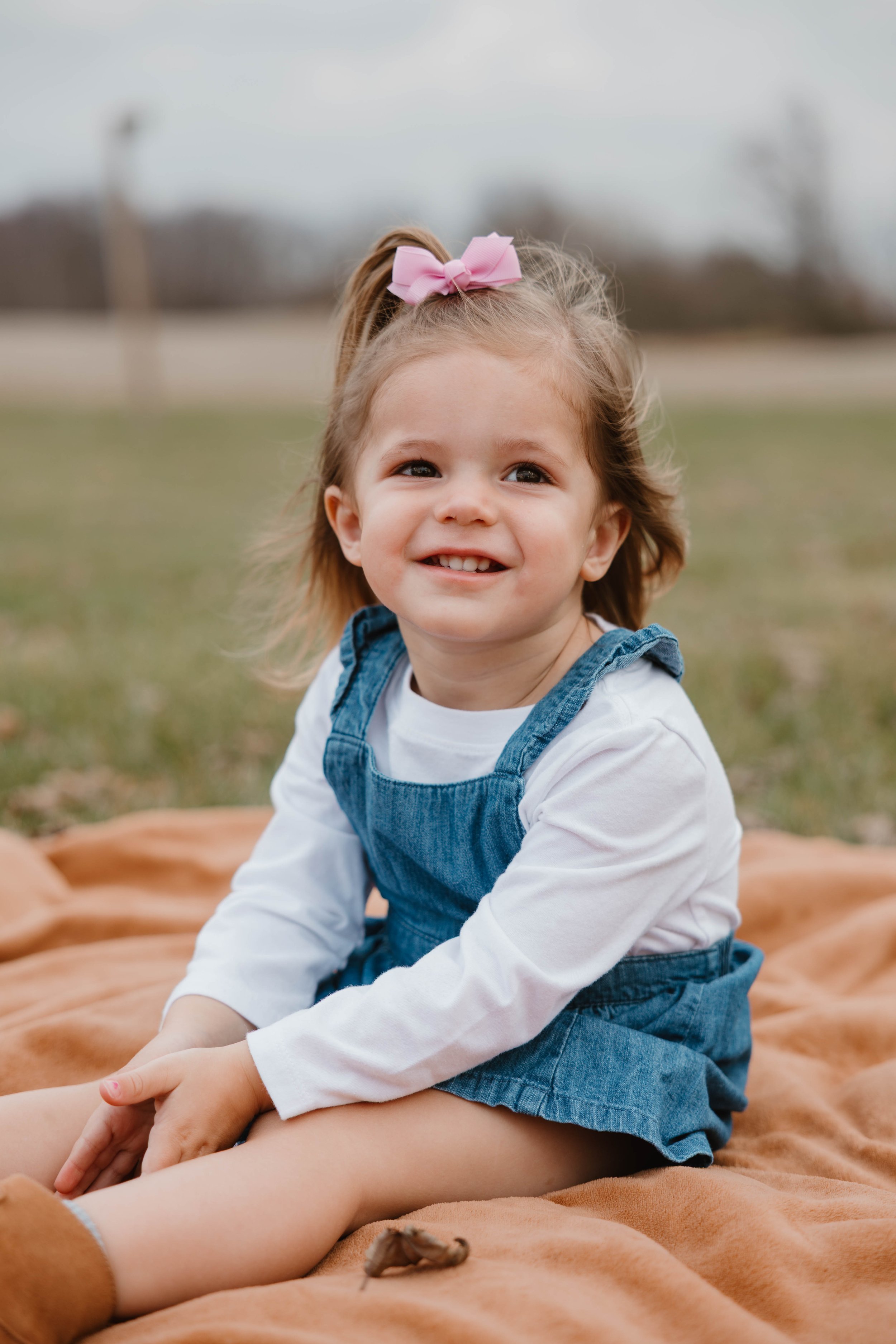 A young girl sitting on a tan blanket outdoors, smiling, wearing a white long sleeve shirt, denim overalls, and a pink bow in her hair, with blurred trees in the background.