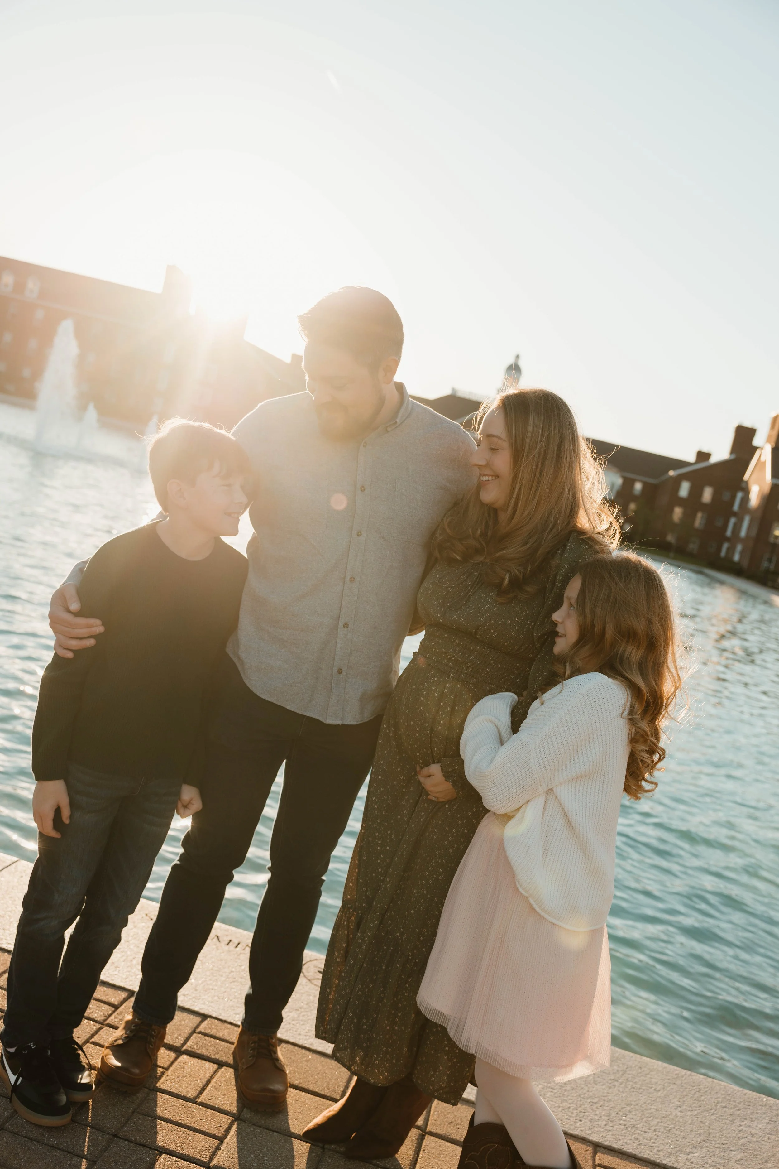 Family of four standing together outdoors near a body of water, smiling, with the sun setting behind them.