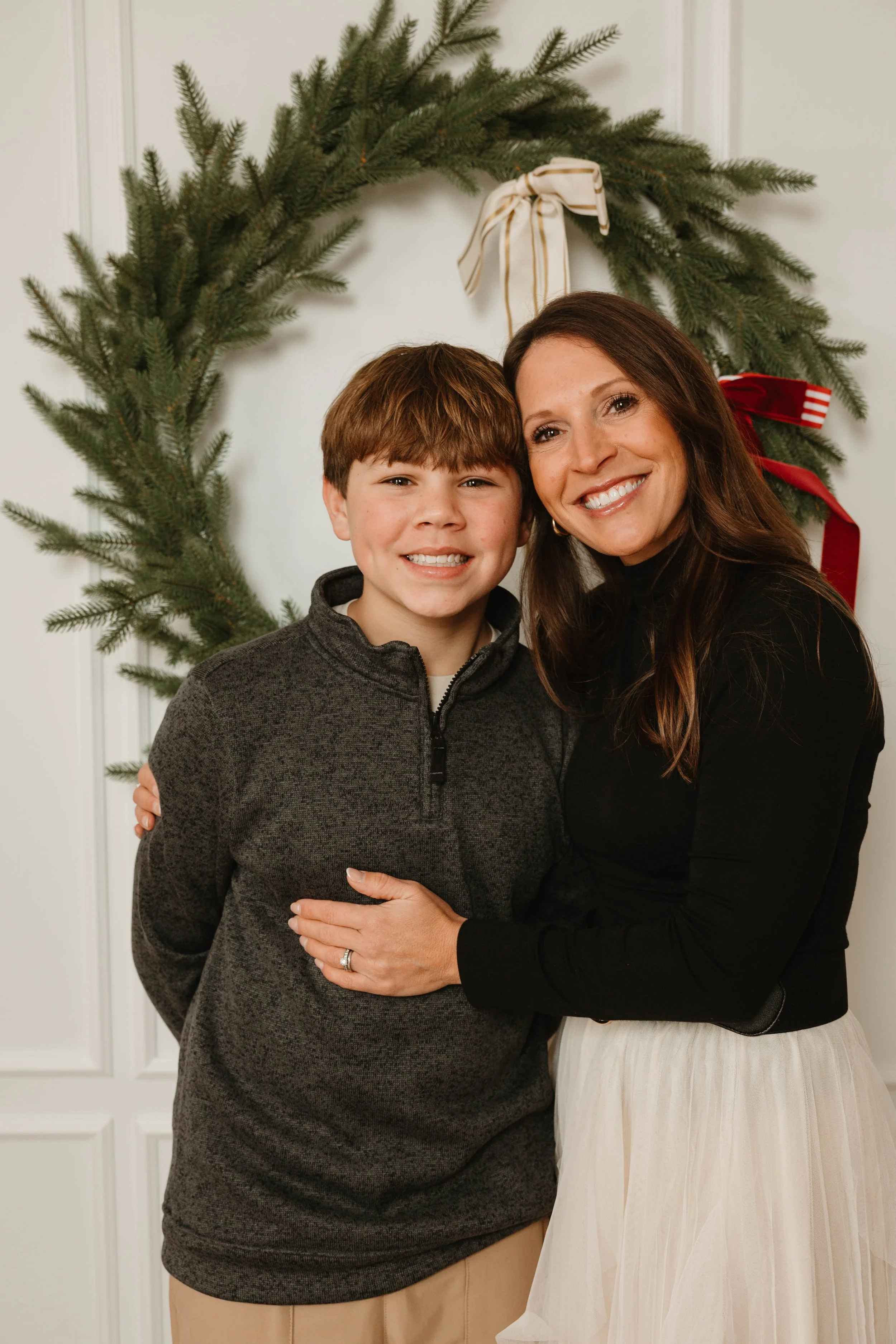 A woman and a boy posing in front of a Christmas wreath with red and white ribbons.