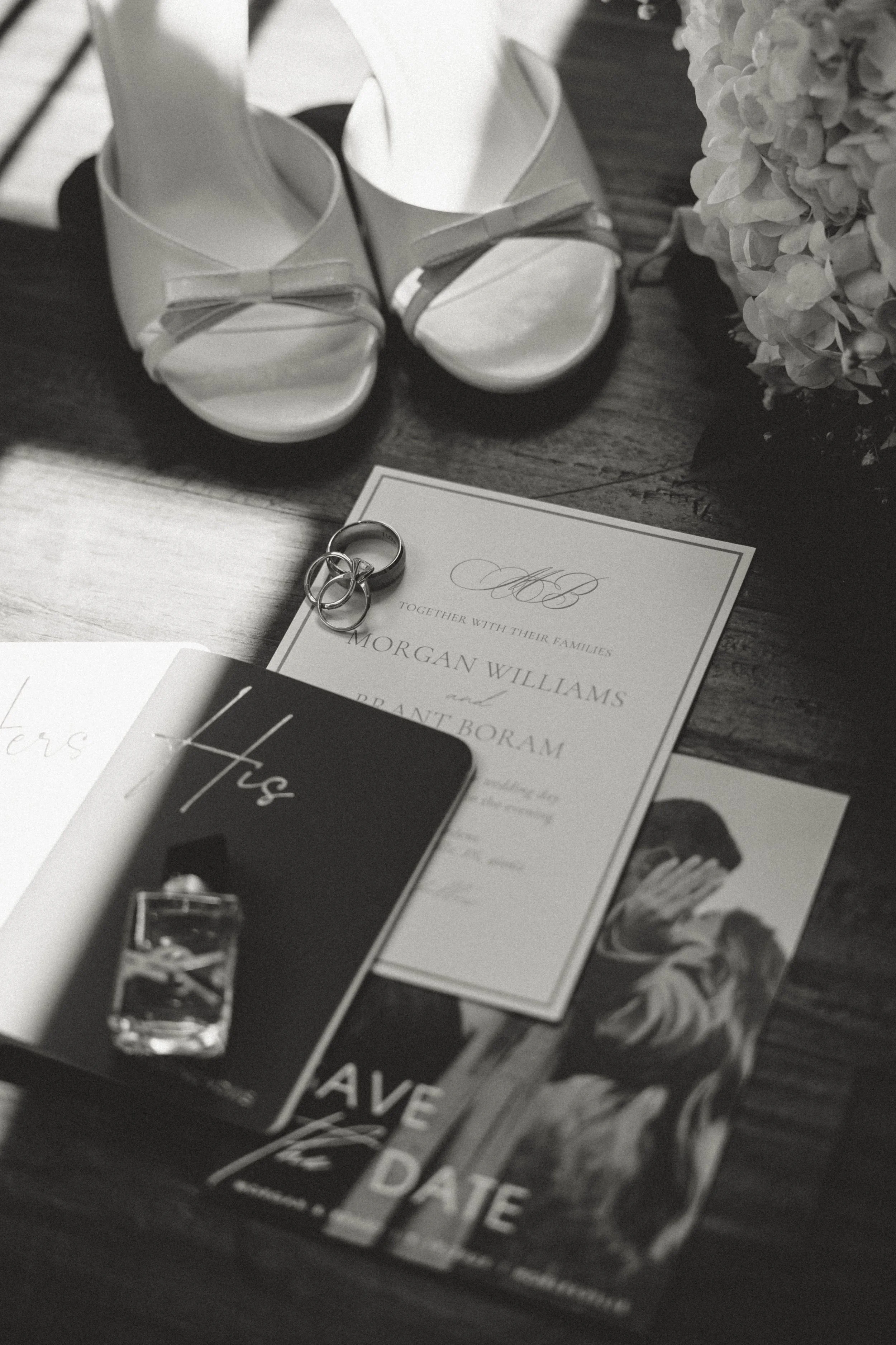 Black and white photo of wedding items including white open-toe heels, wedding rings, a wedding invitation for Morgan Williams and Grant Boram, a guidebook with a ring photo, a brochure with a couple and "SAVE THE DATE" text, and a floral arrangement