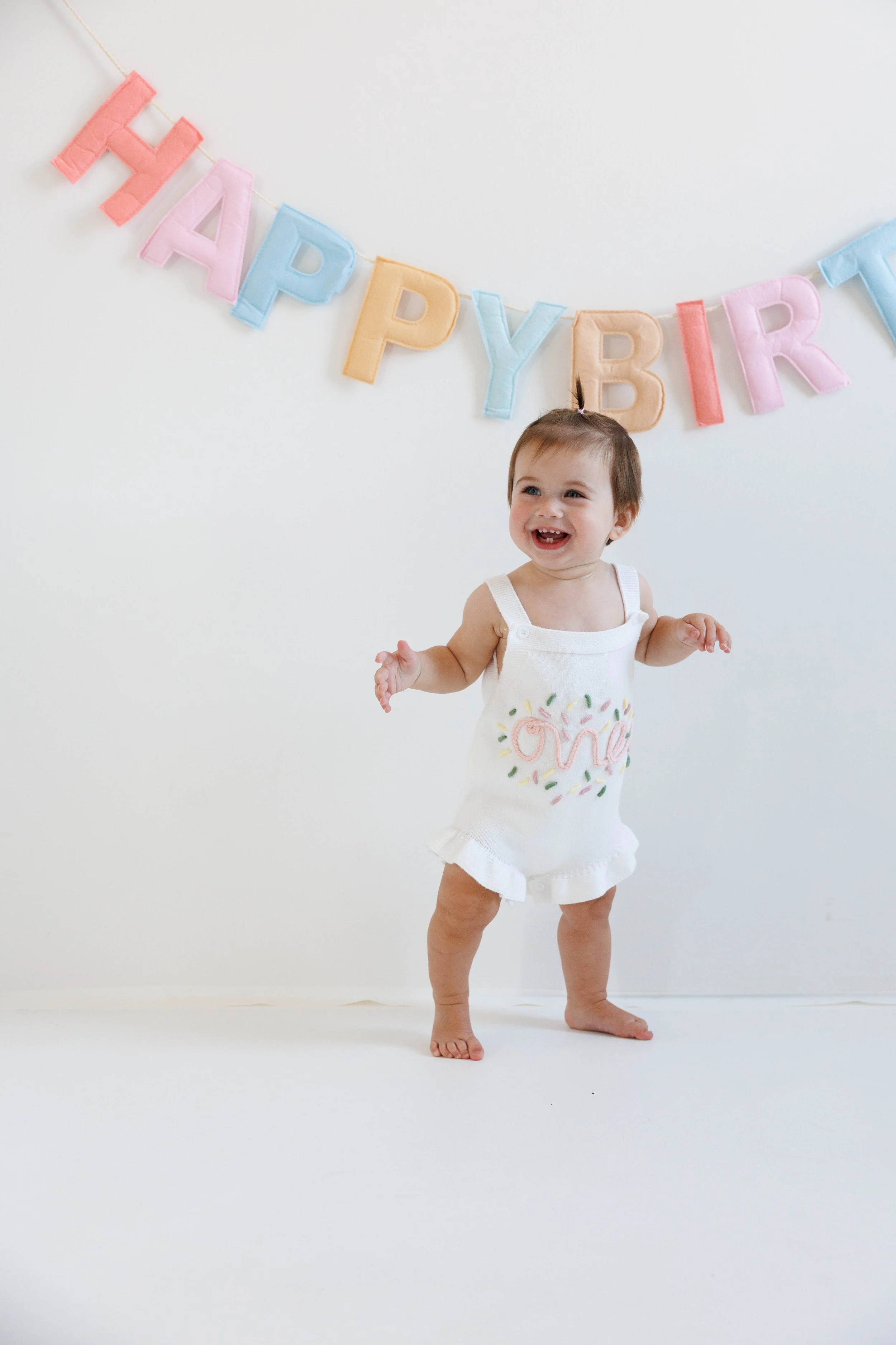 A smiling toddler girl in a white romper with "one" embroidered on it, standing on a white floor against a plain white wall, with a pastel-colored "Happy Birthday" banner hanging above her.