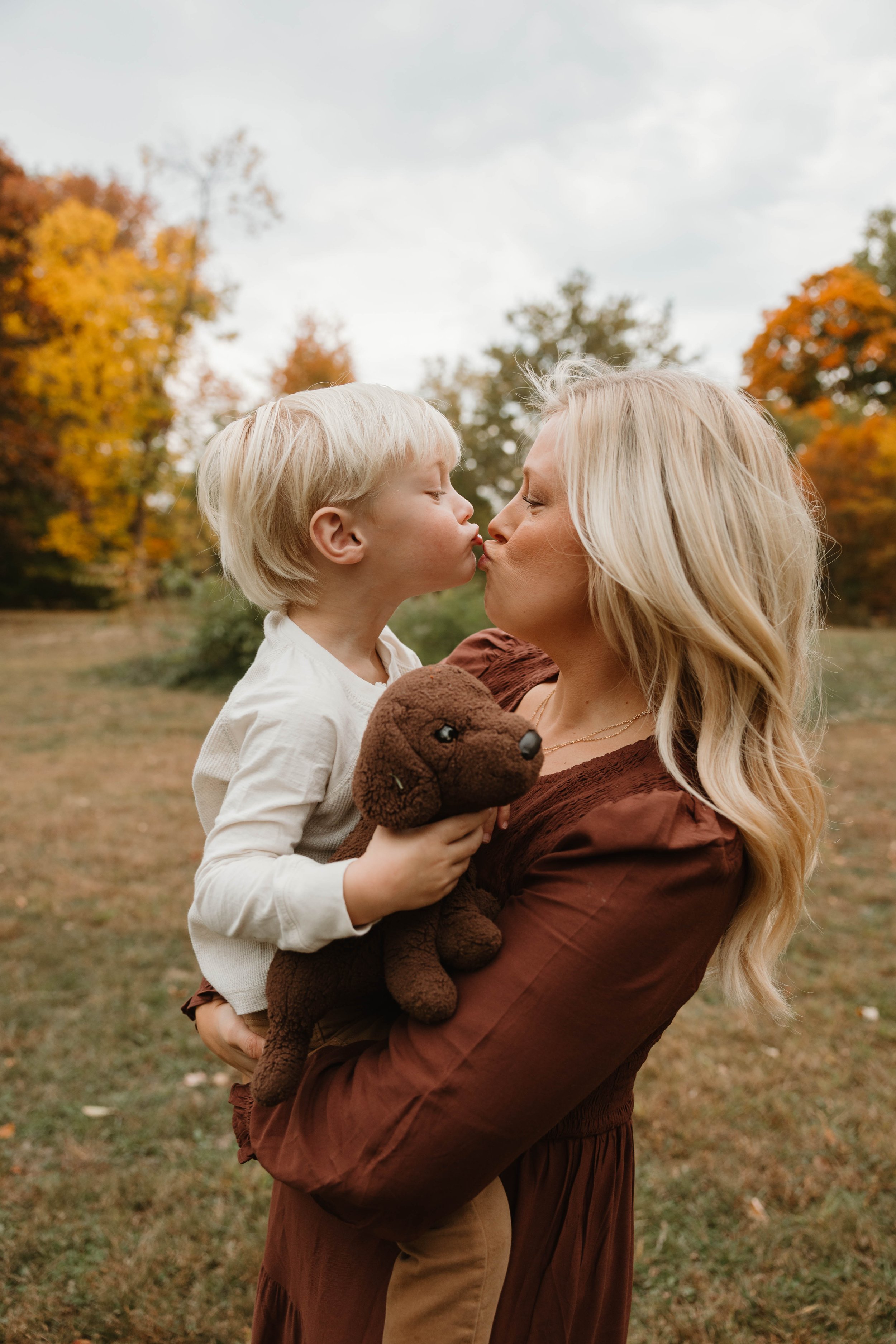 A woman holding a young boy outdoors on a fall day, about to kiss him. The boy is holding a brown stuffed dog, and the background features trees with orange and yellow leaves.