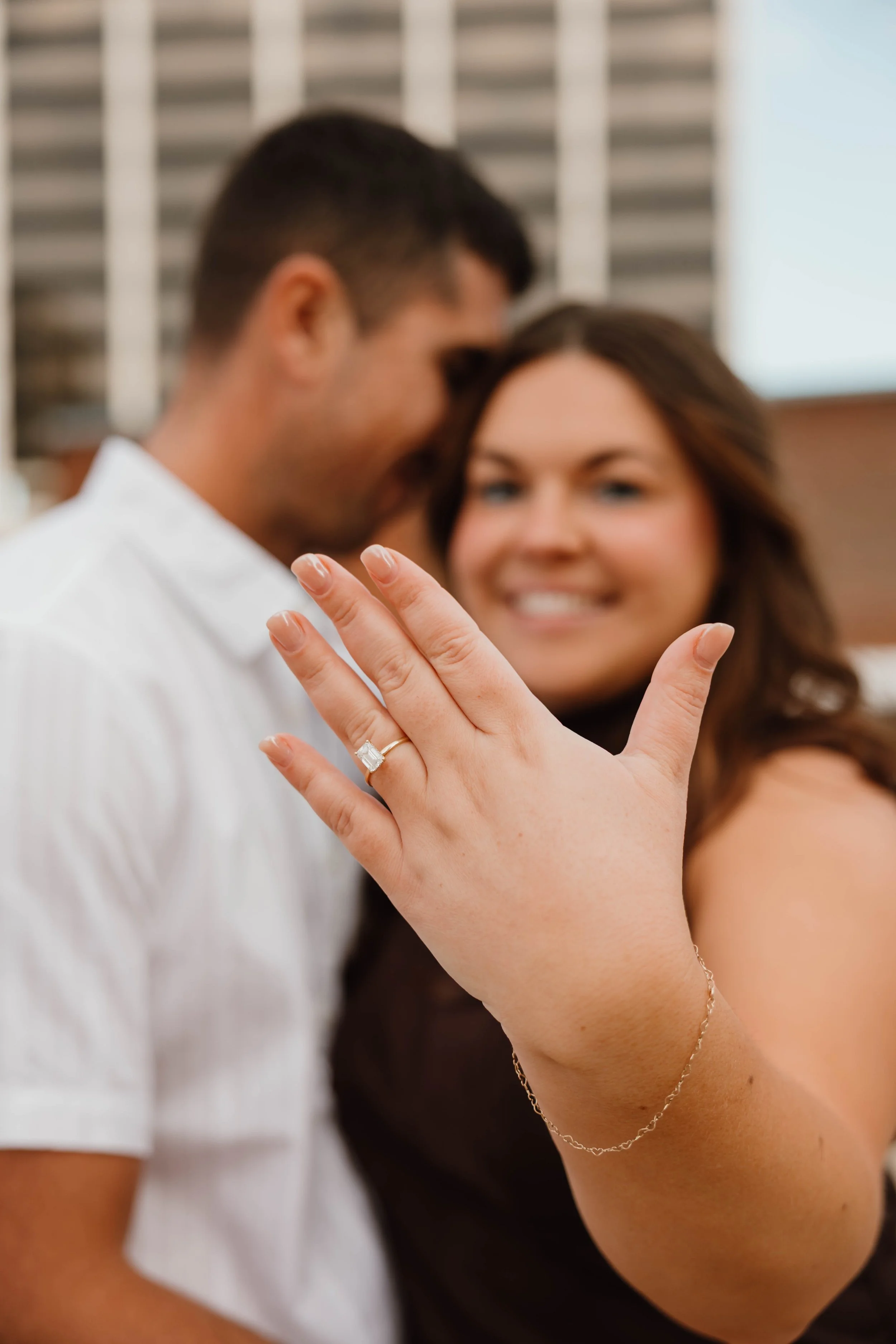 Woman showing her engagement ring on her left hand in front of smiling man and woman with blurred background.