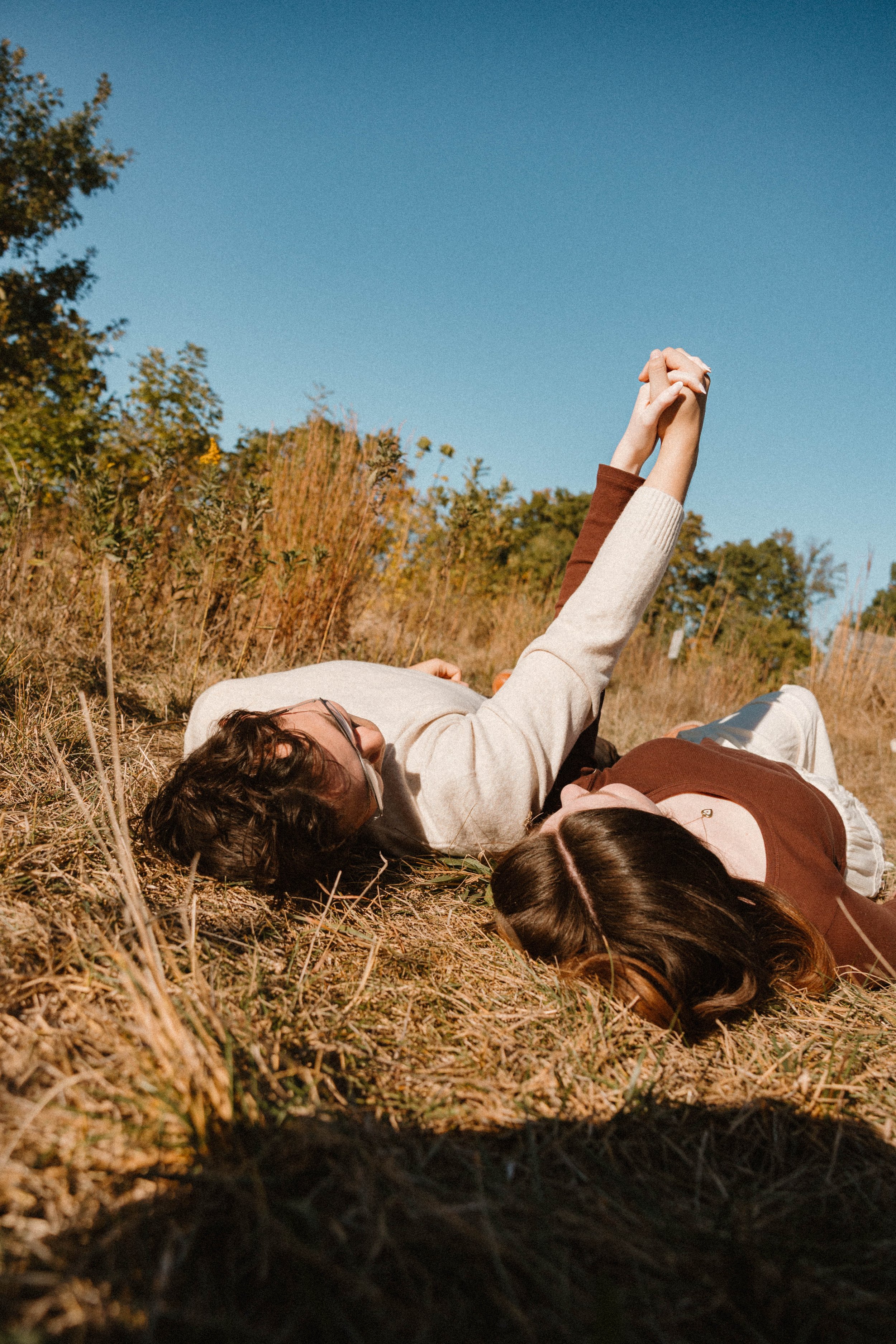 A couple lying on the grass outdoors on a sunny day, holding hands and looking at each other.