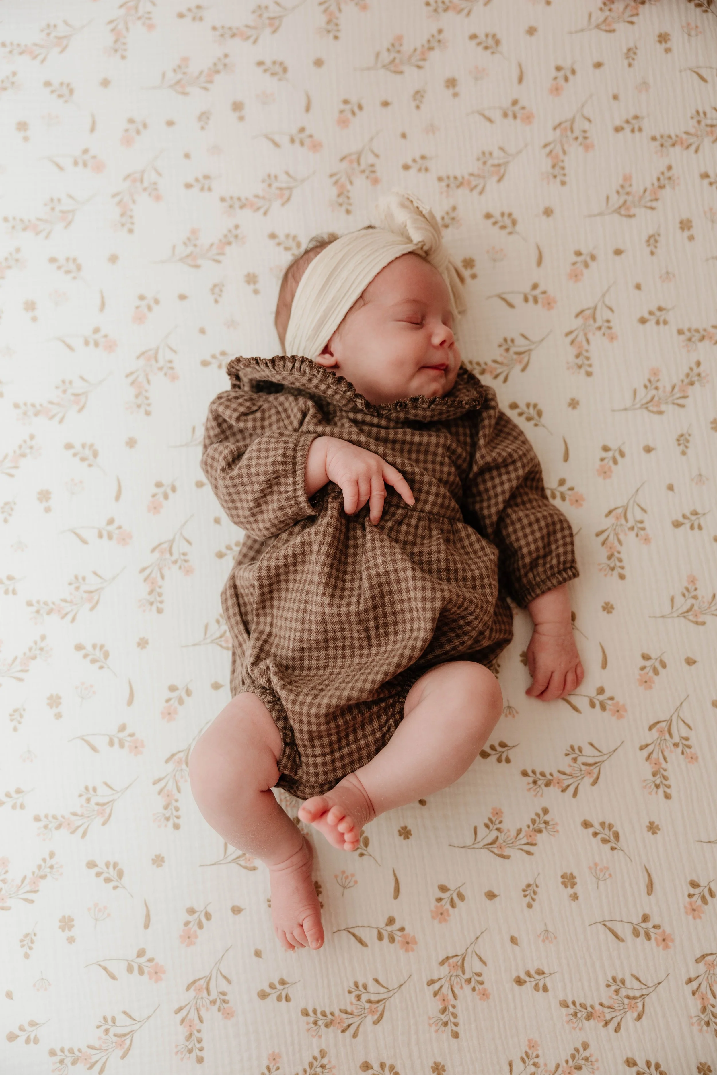 A newborn baby is sleeping on a bed with a floral patterned sheet, wearing a brown checkered outfit and a cream-colored headband.