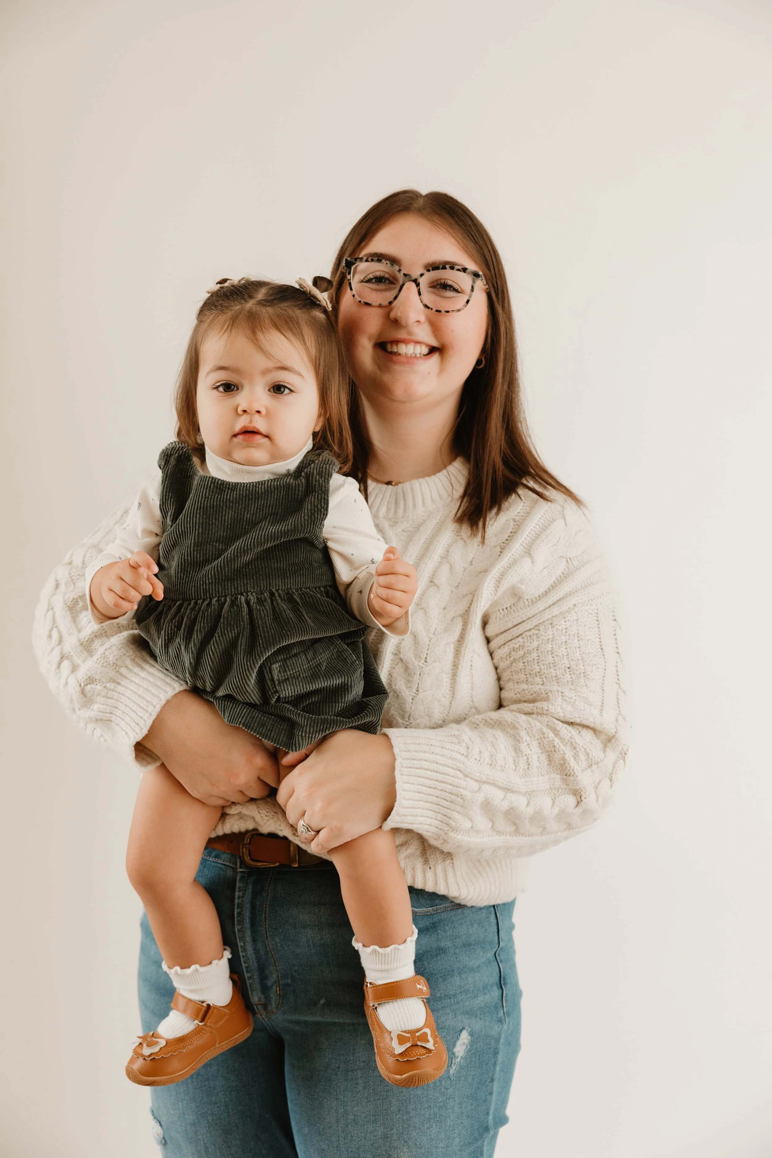 A smiling woman with glasses holding a young girl in front of a plain white background.