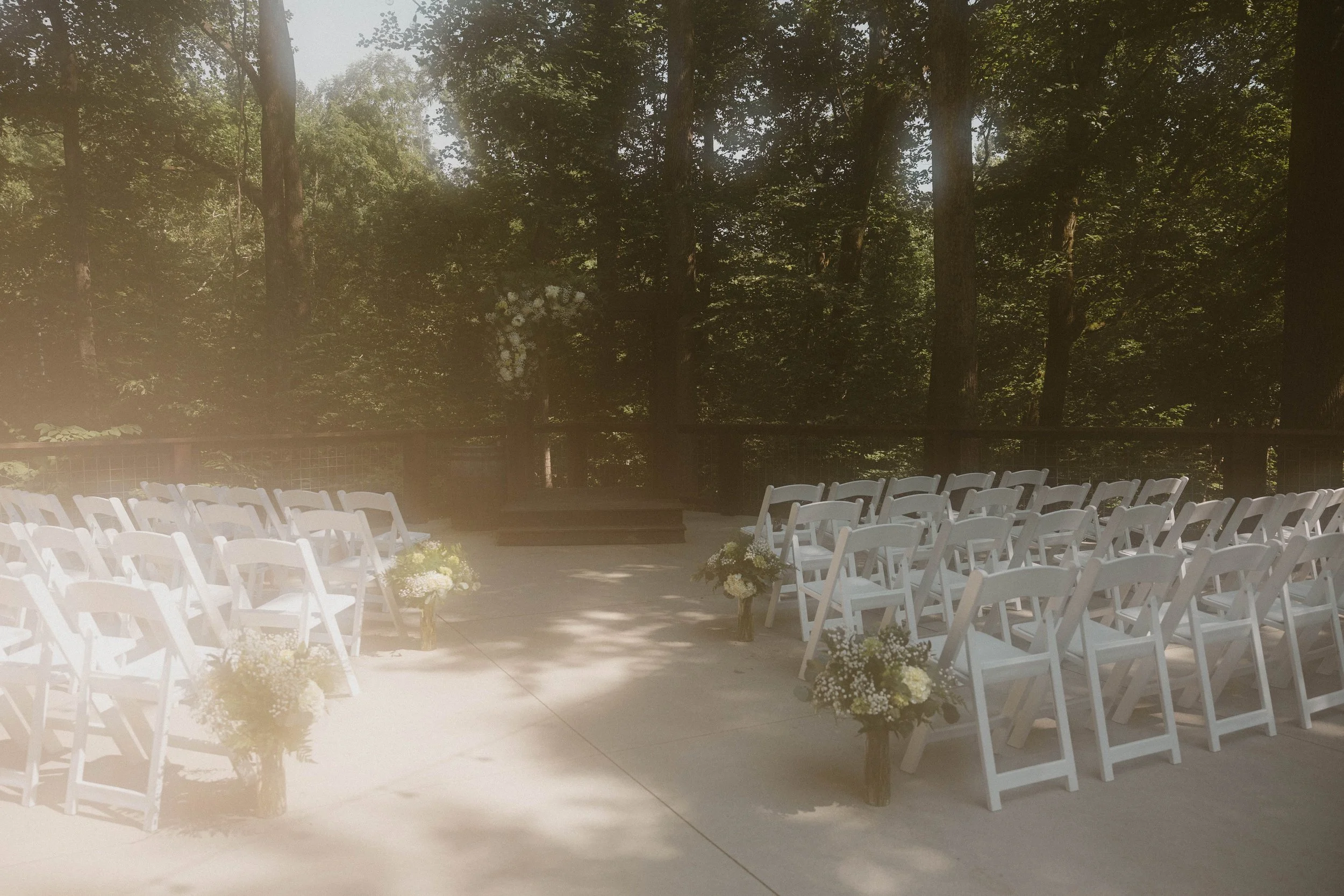 Empty outdoor wedding ceremony setup with white chairs and flower arrangements on a patio surrounded by trees.
