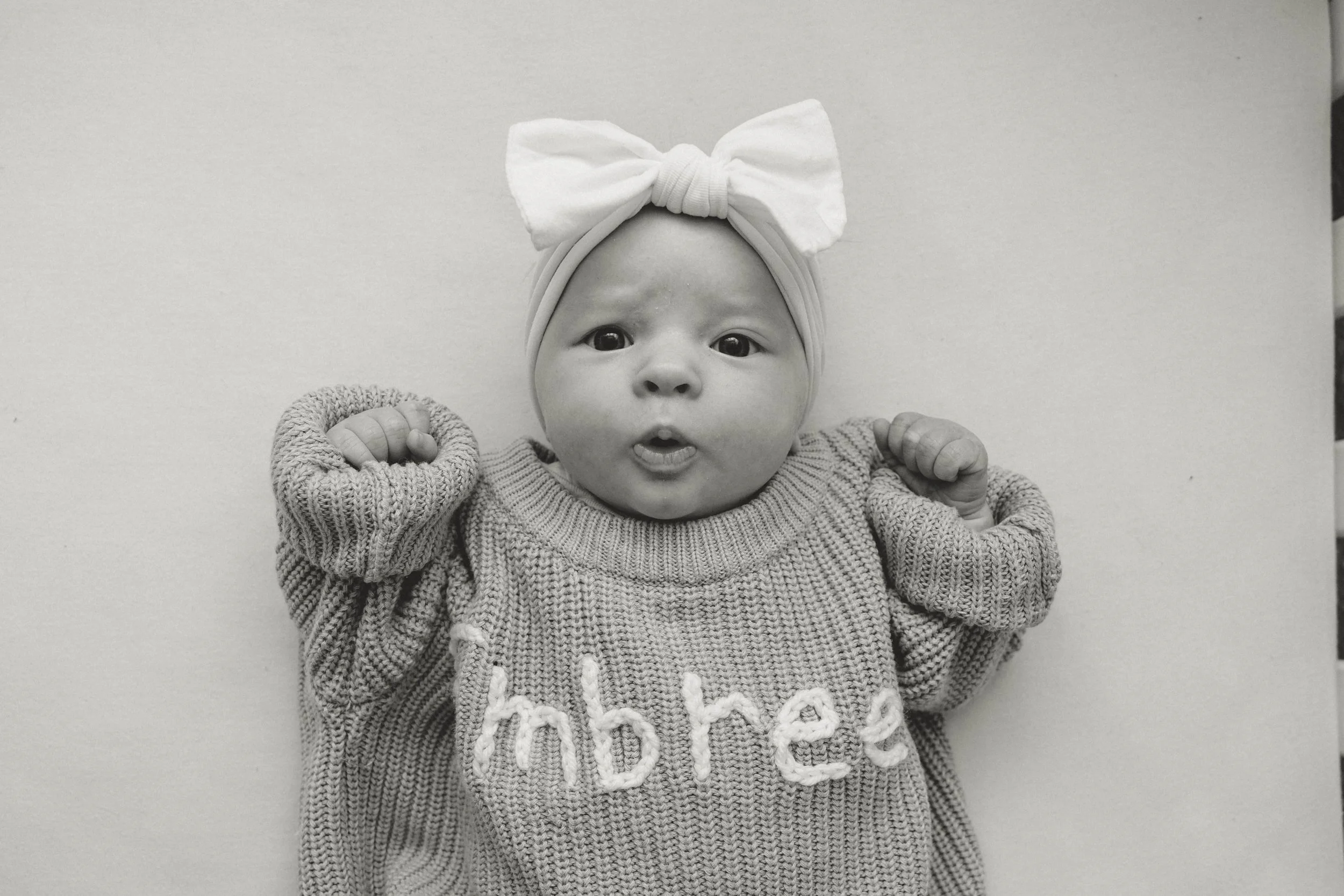 Baby wearing a headband with a bow, holding fists up, wearing a sweater with the word 'embree' on it.