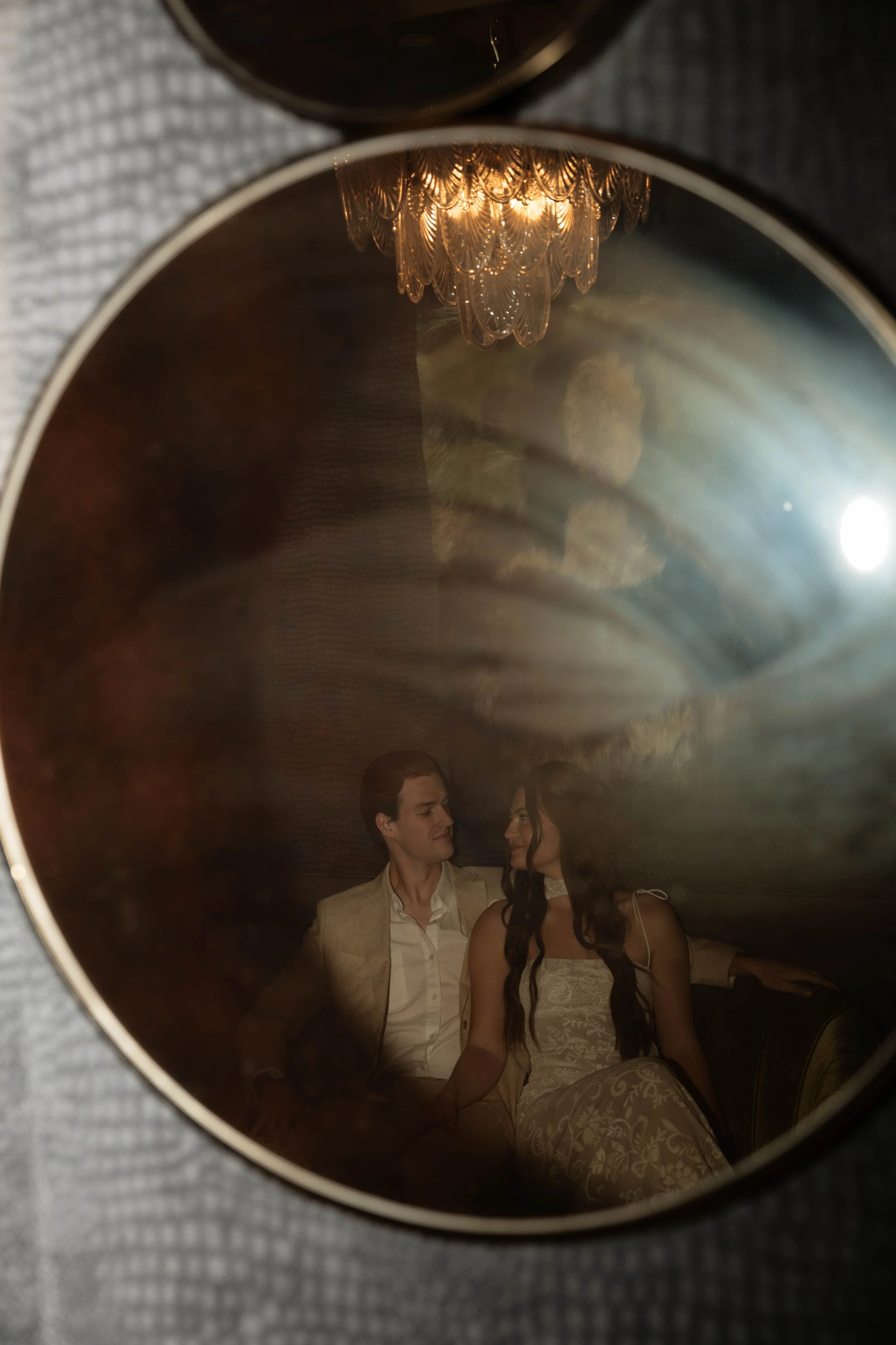 A couple sitting closely inside a reflective surface showing a chandelier above them, with the woman wearing a lace dress and the man in a beige suit.