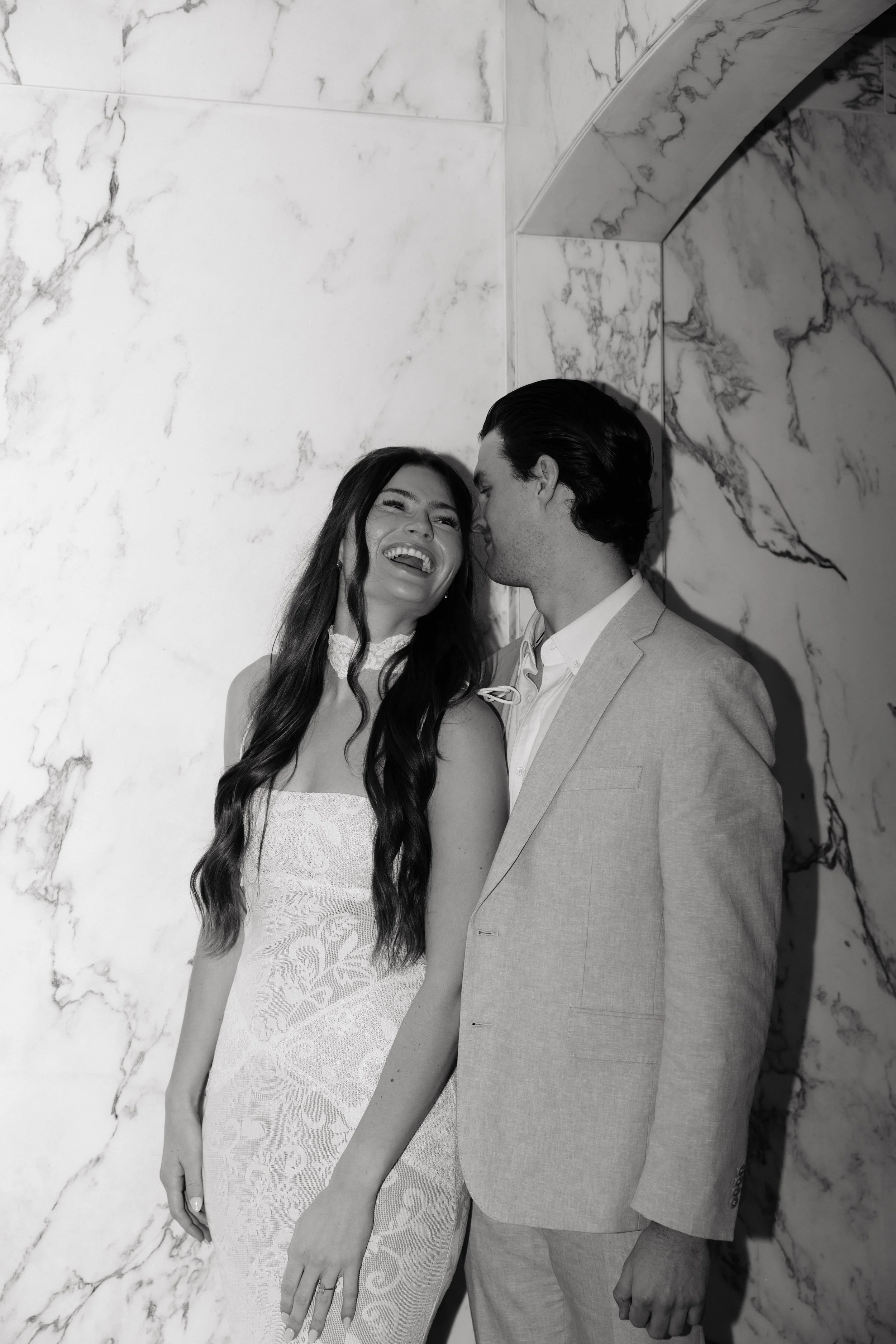 A Black and White photo of a smiling woman in a lace dress and a man in a light suit sharing an intimate moment, with the woman laughing and the man leaning towards her, against a marble wall background.