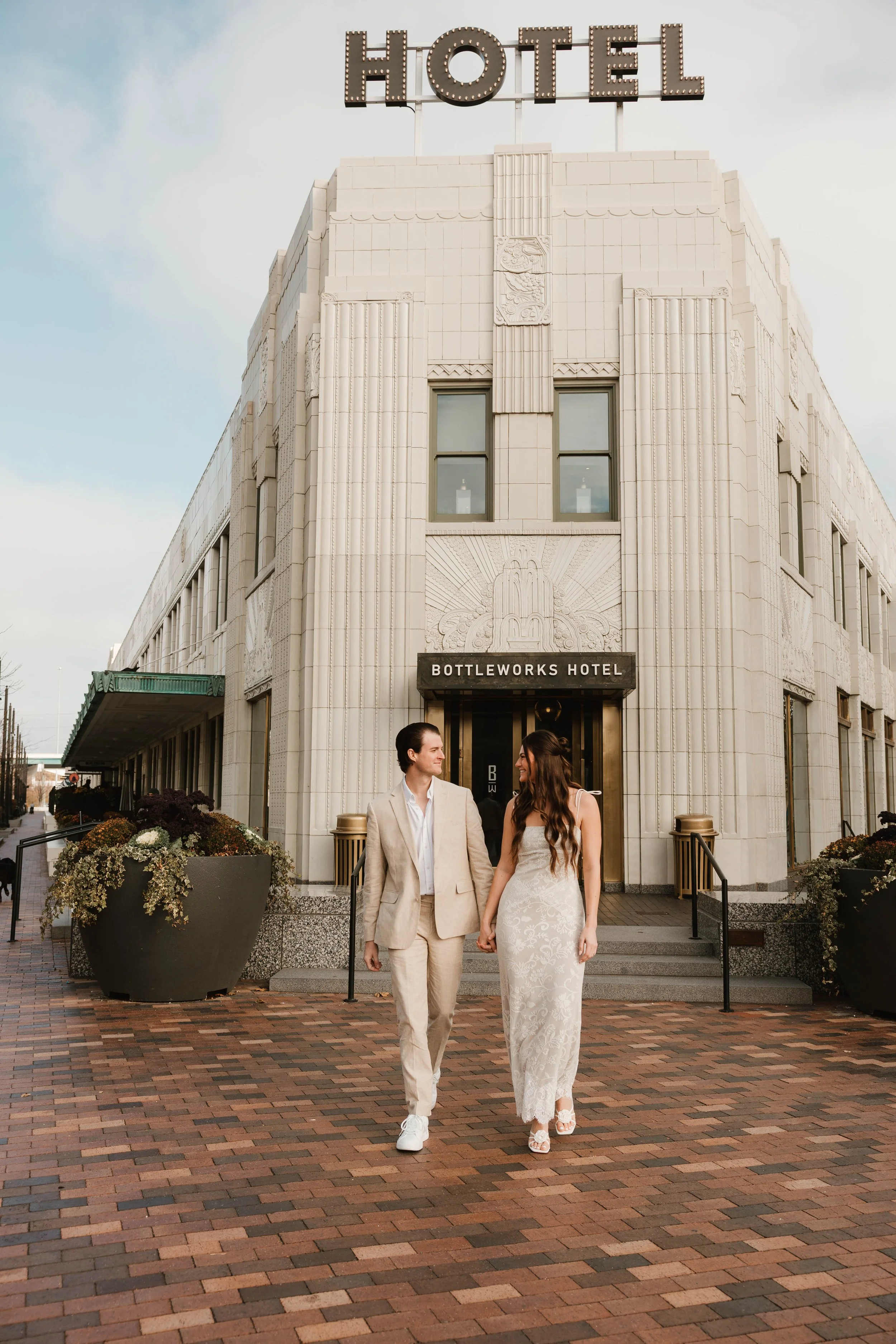 A man and woman in formal attire holding hands and walking out of the Bottleworks Hotel building, with a large illuminated 'HOTEL' sign on top.