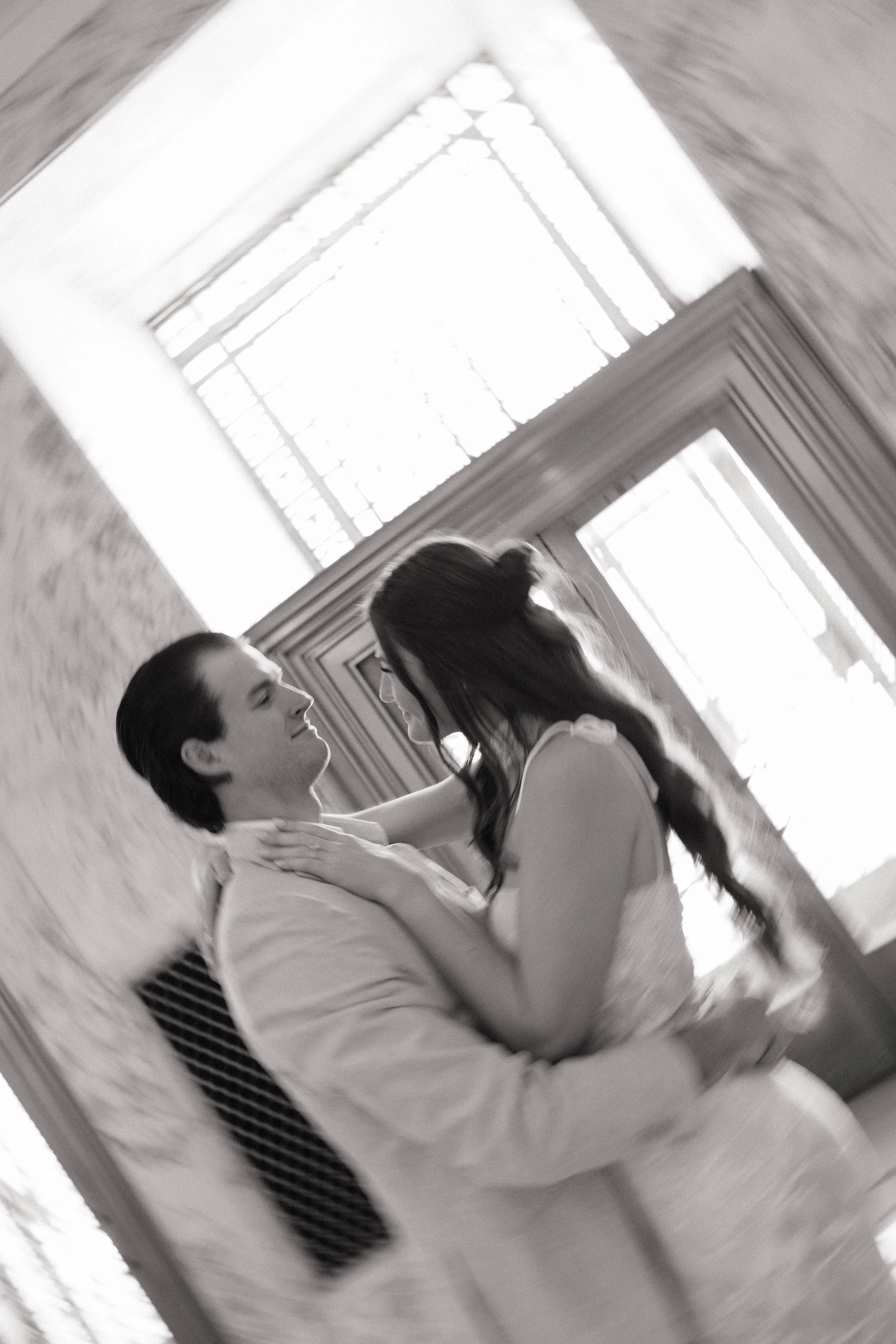 A black and white photo of a couple dancing indoors, with large windows and wooden paneling in the background.