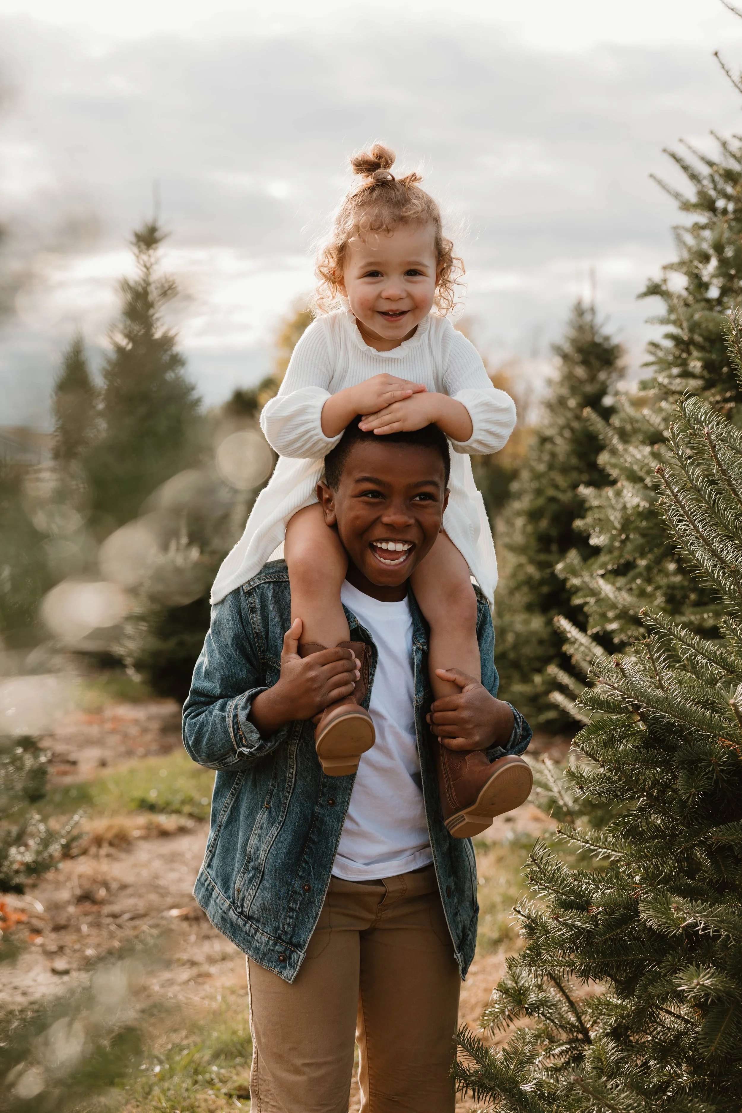 A young man carrying a small girl on his shoulders among Christmas trees outdoors on a cloudy day.