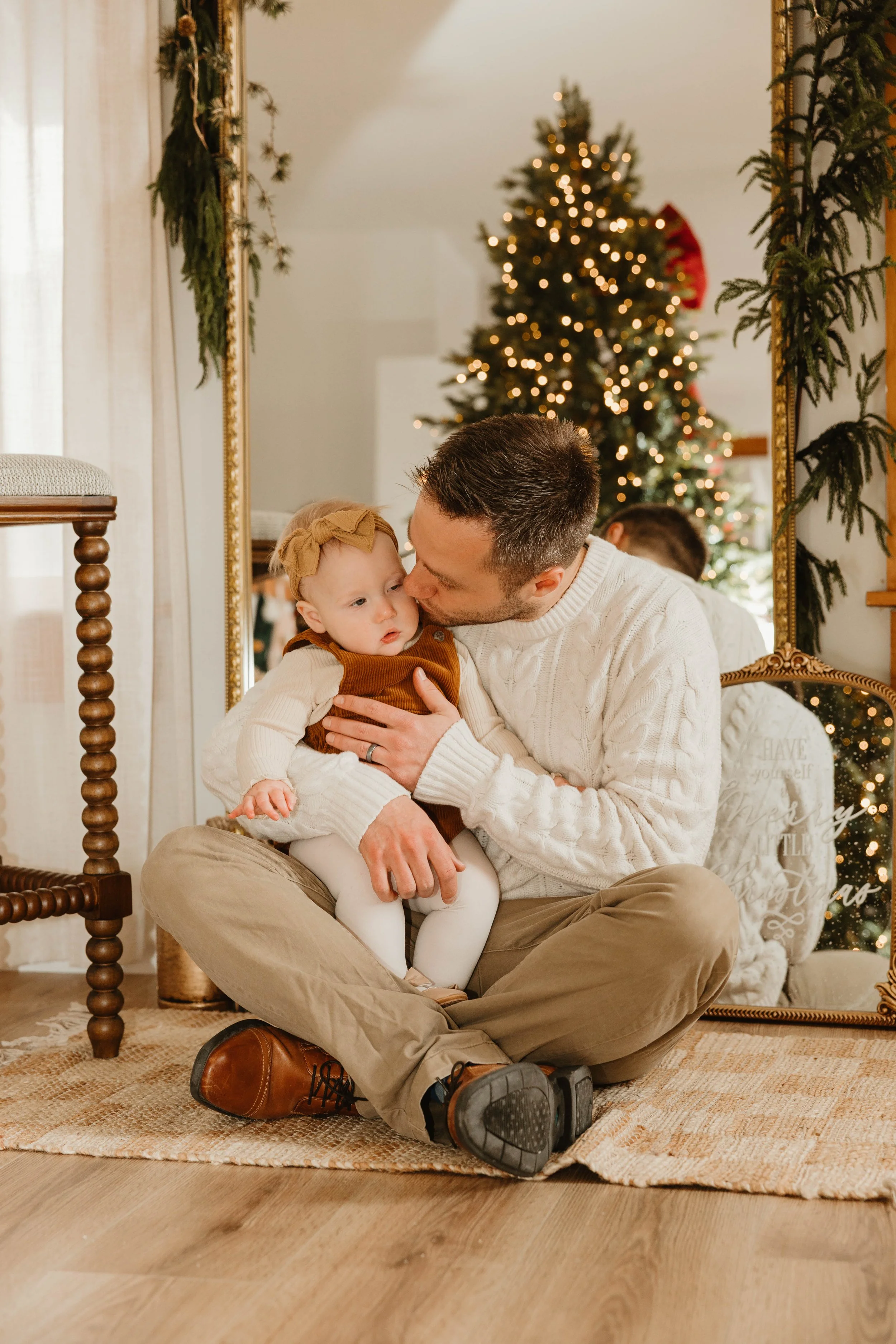 A man with short dark hair, wearing a white sweater and tan pants, sitting cross-legged on a rug on a wooden floor, holding and kissing a young girl with light hair, wearing a brown dress, white tights, and a beige headband. They are in a room decora
