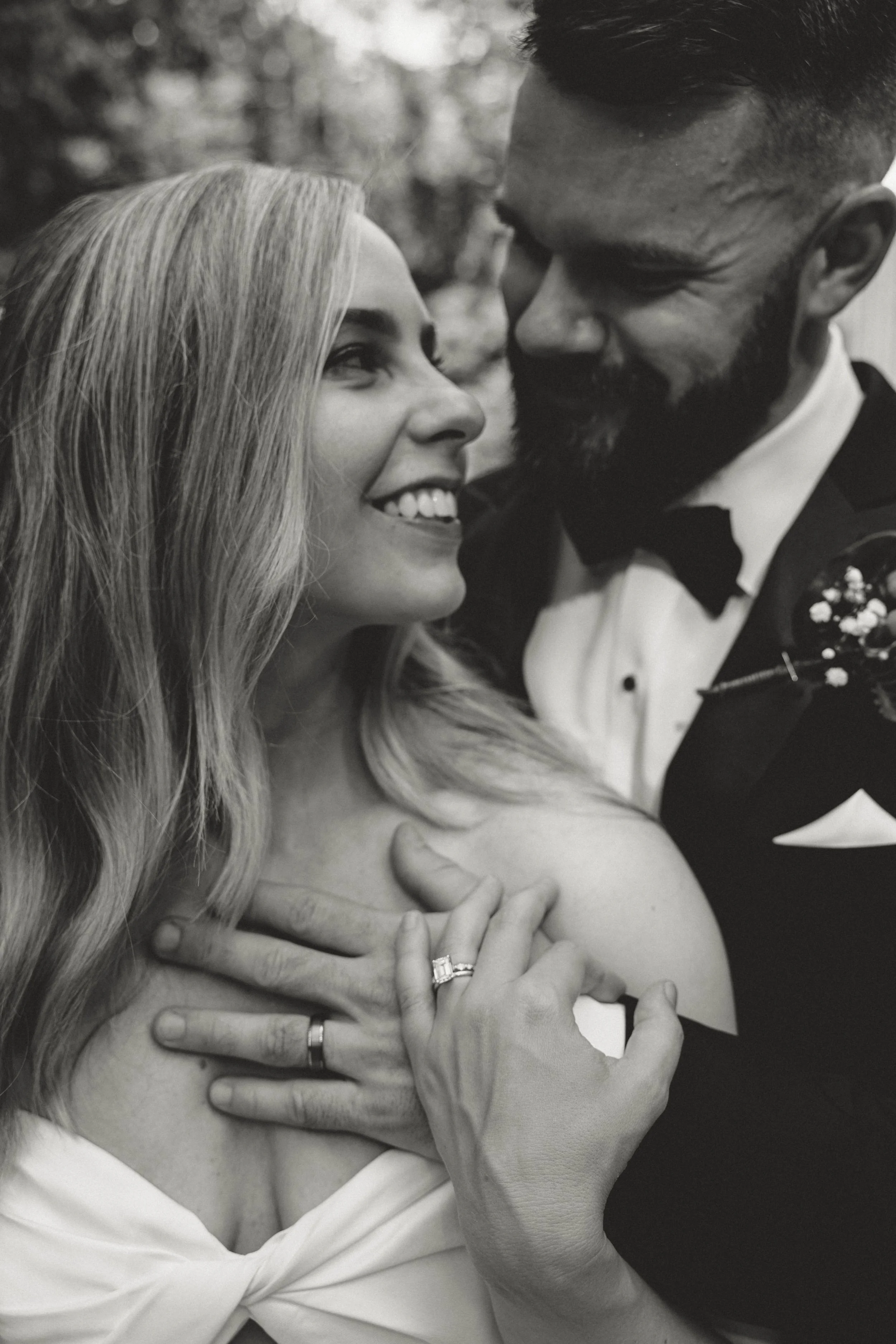 A black and white photo of a happy couple on their wedding day, embracing and smiling at each other. The woman shows her wedding ring, and they are dressed in formal wedding attire.