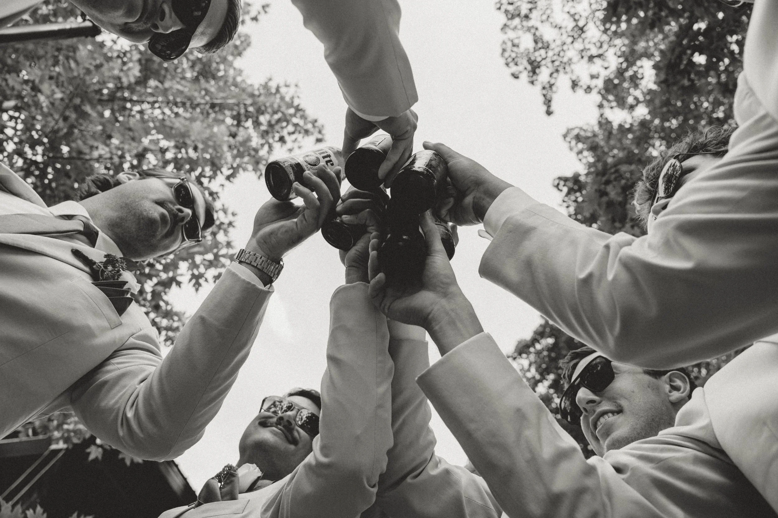 A group of men in suits and sunglasses are raising their bottles in a toast, viewed from below with trees and sky in the background.