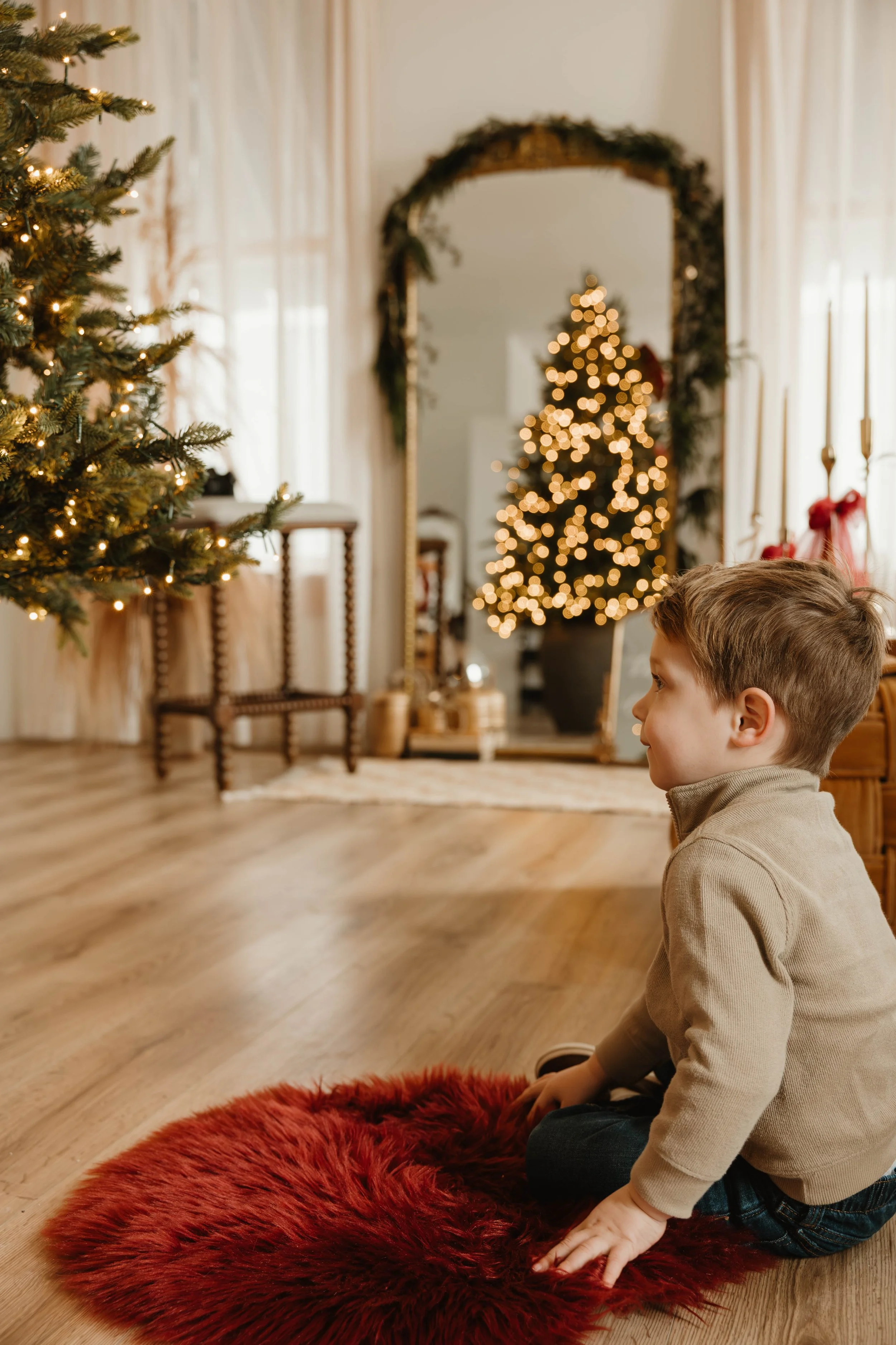 A young boy sitting on a red furry rug on the wooden floor, looking at a decorated Christmas tree with lights. In the background, a large mirror with a garland frame reflects a second, decorated Christmas tree with lights.