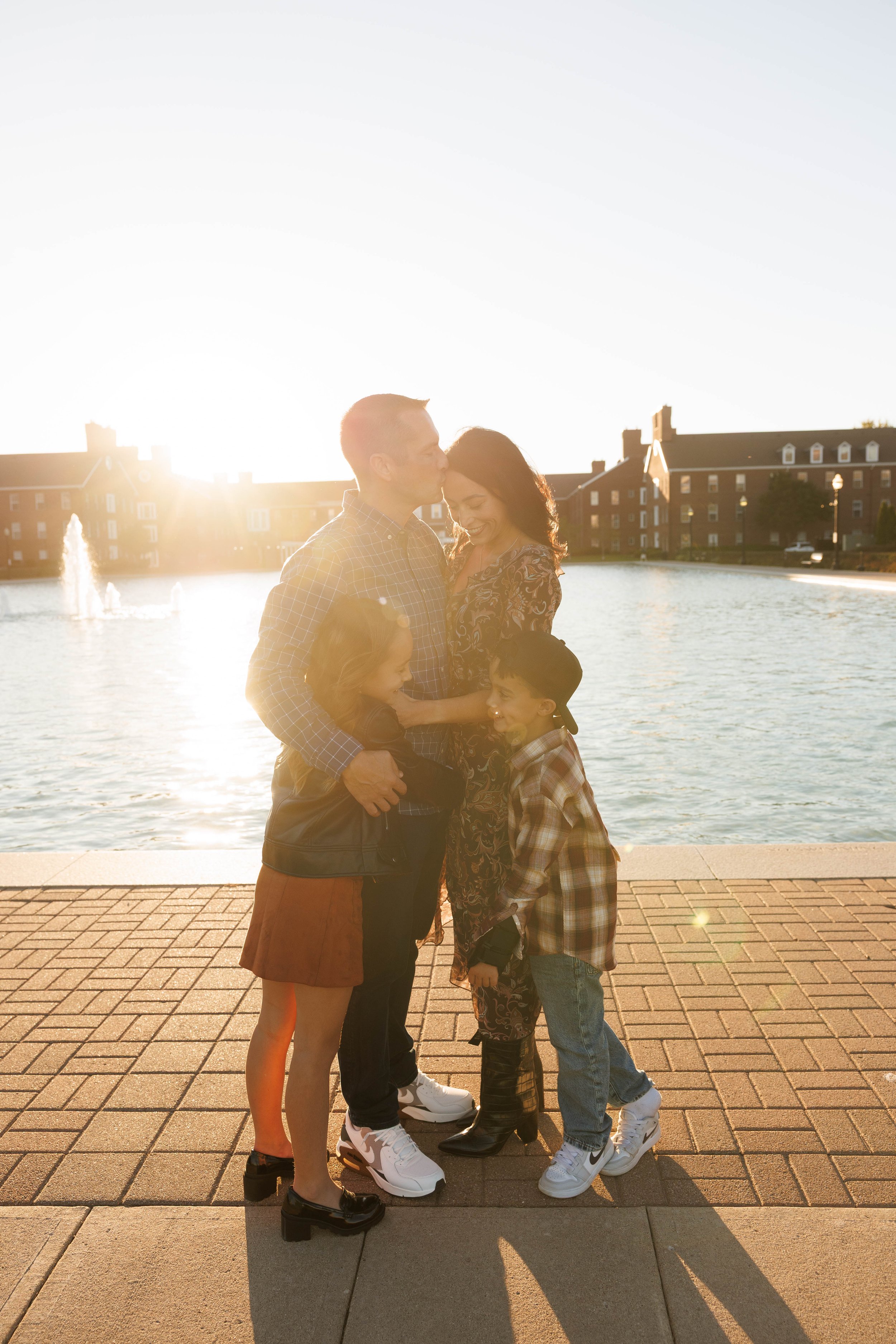 A happy family of four standing by a lakeside with fountains, during sunset, with brick buildings in the background. The parents are kissing their children, who are smiling.
