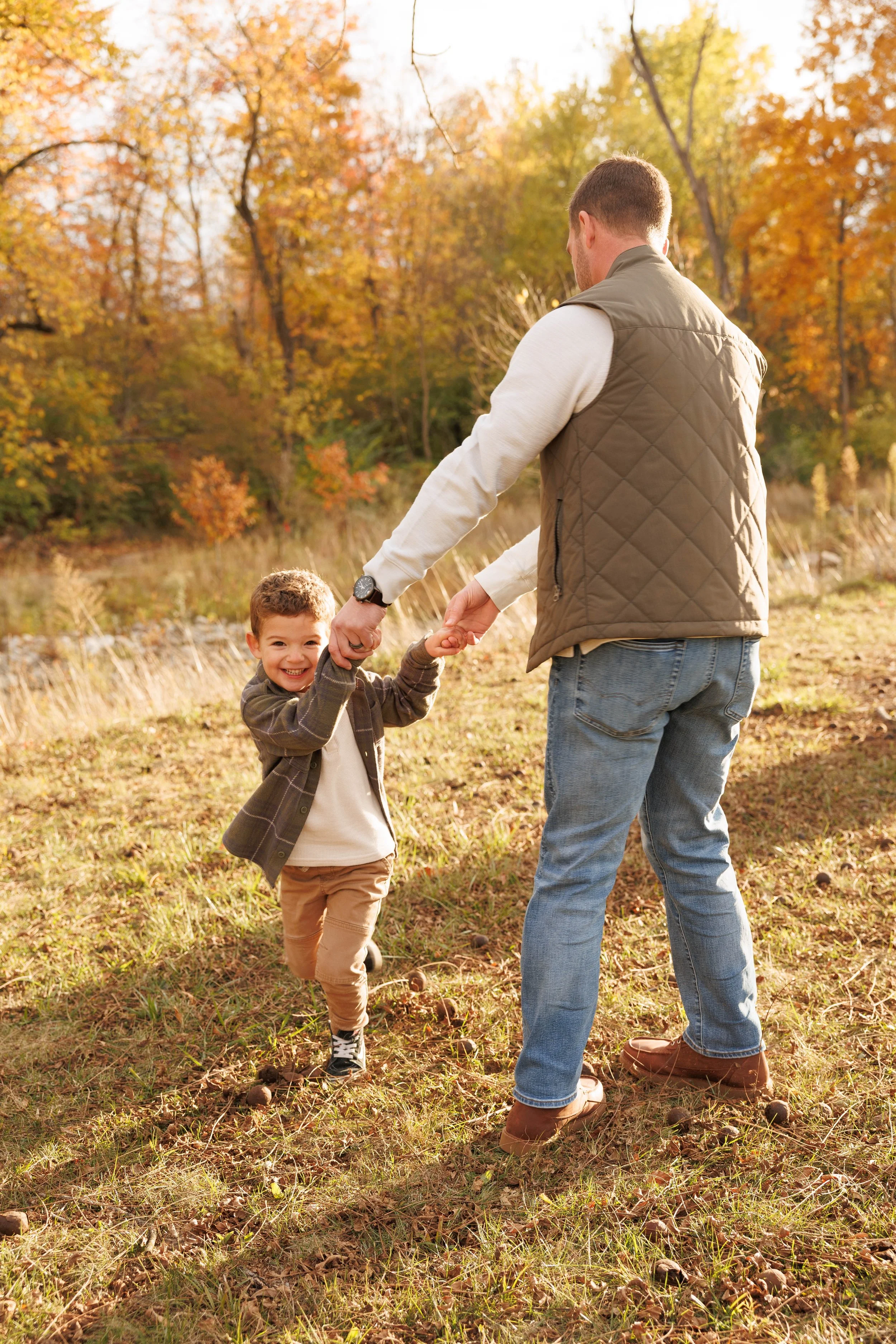 A man and a young boy are holding hands and playing outdoors in a grassy, wooded area during fall. The boy is smiling and looking at the camera, while the man is turned away.