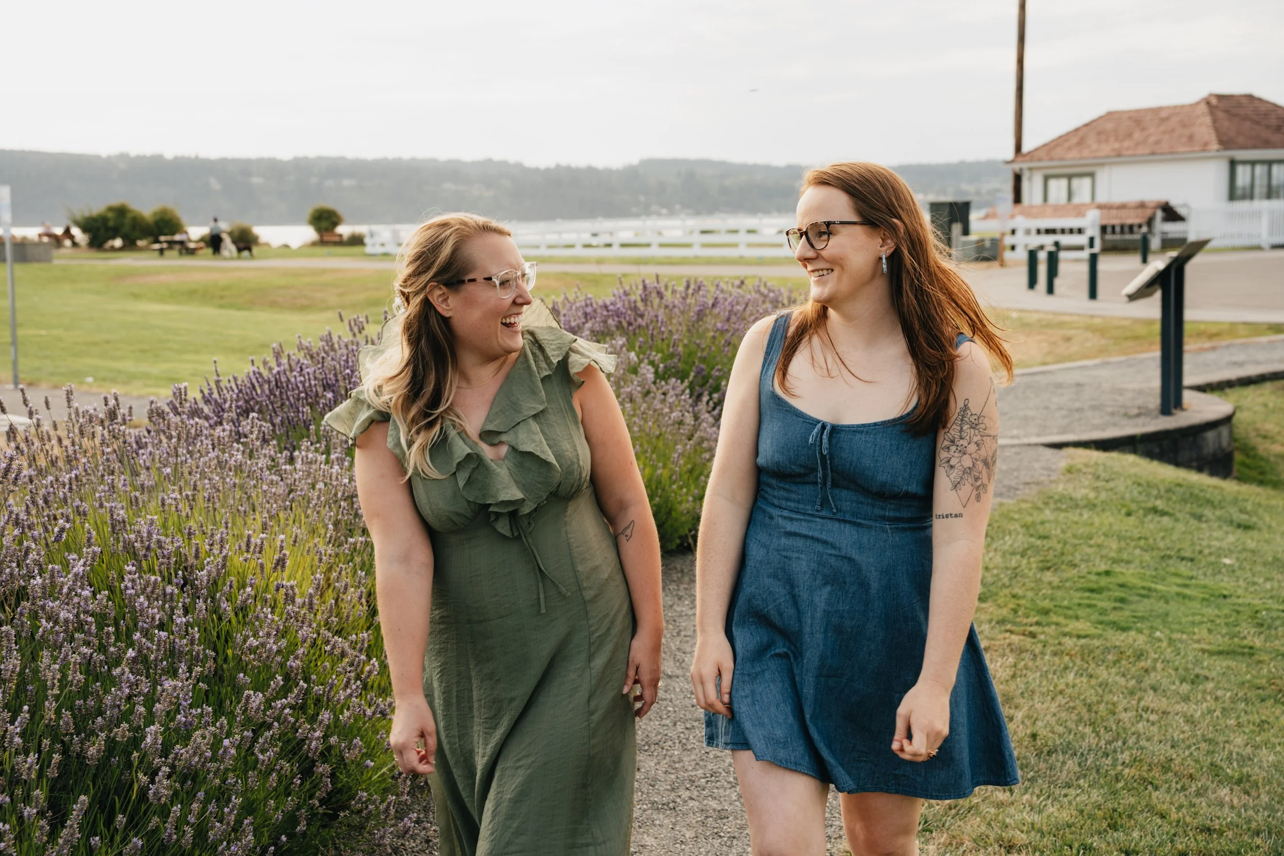Two women smiling and walking outdoors near a lavender flower bed with a body of water and buildings in the background.