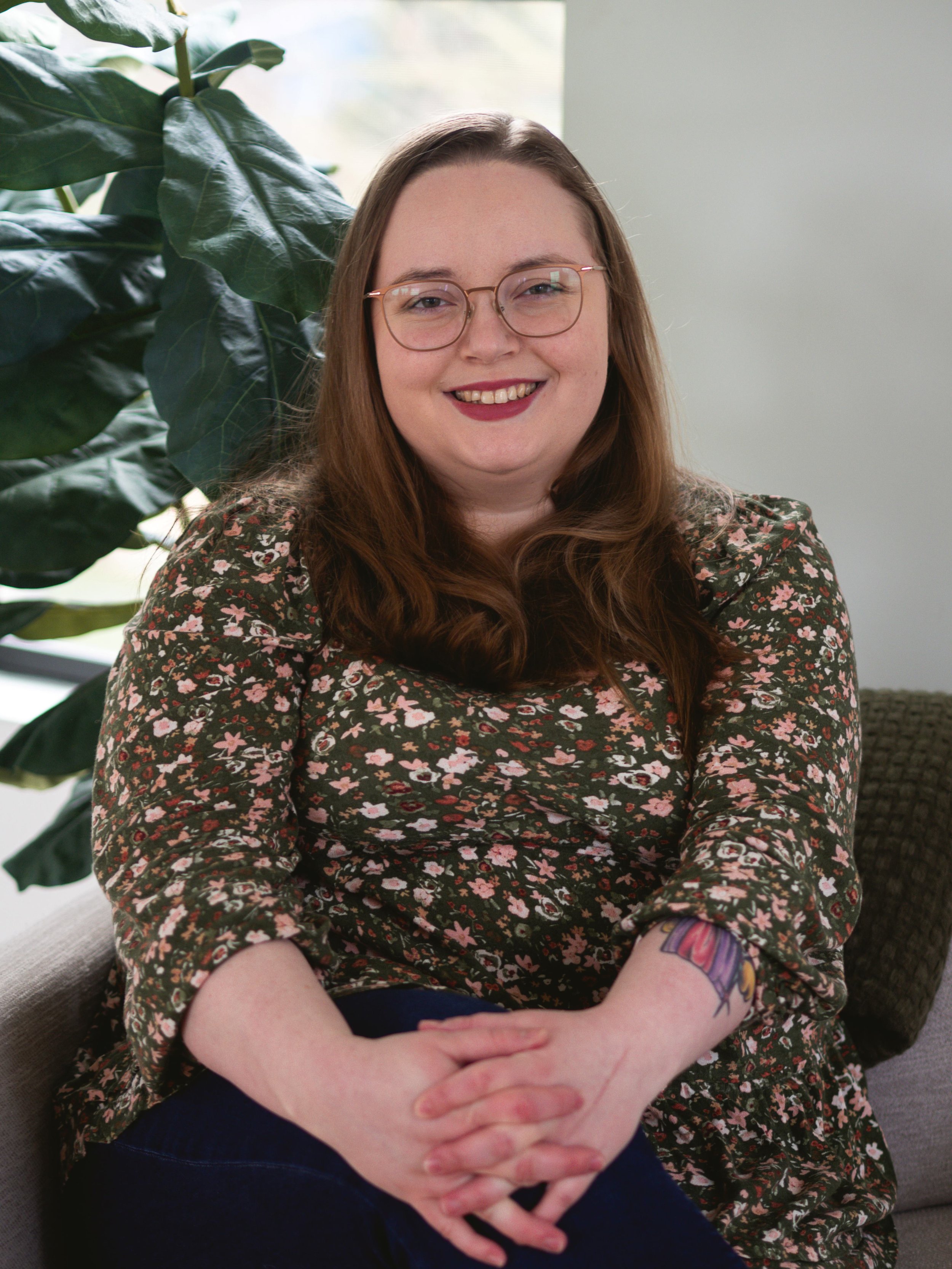 A smiling woman with long red hair, glasses, and a floral dress sitting indoors with a green plant behind her and a white wall to her right.