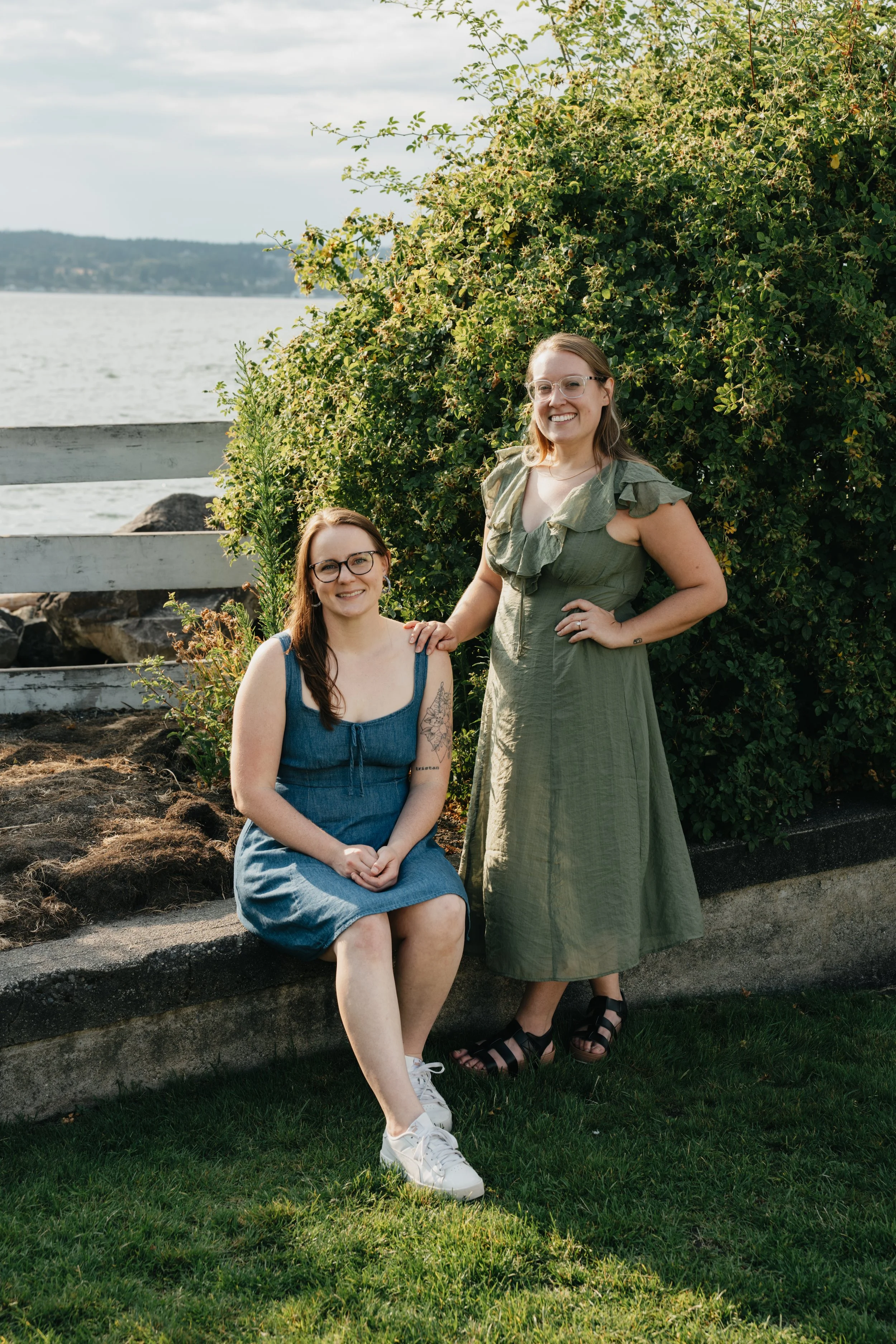 Two women posing outdoors near a large bush with a body of water in the background. One woman is seated on a concrete ledge wearing a blue dress, white sneakers, and glasses. The other woman stands beside her wearing a green dress, black sandals, and glasses, with her hand on the seated woman's shoulder.