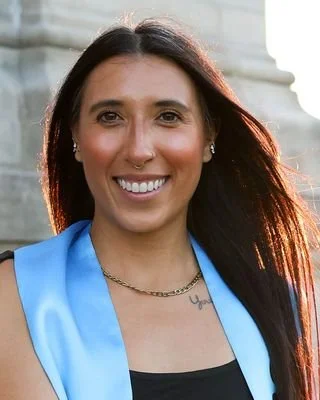 A smiling young woman in a black dress sitting on a stone ledge outside a large stone building at sunset, wearing a blue sash that reads 'Turning the Mental Health Stigma Inside Out' and holding a black bag with various health-themed stickers.