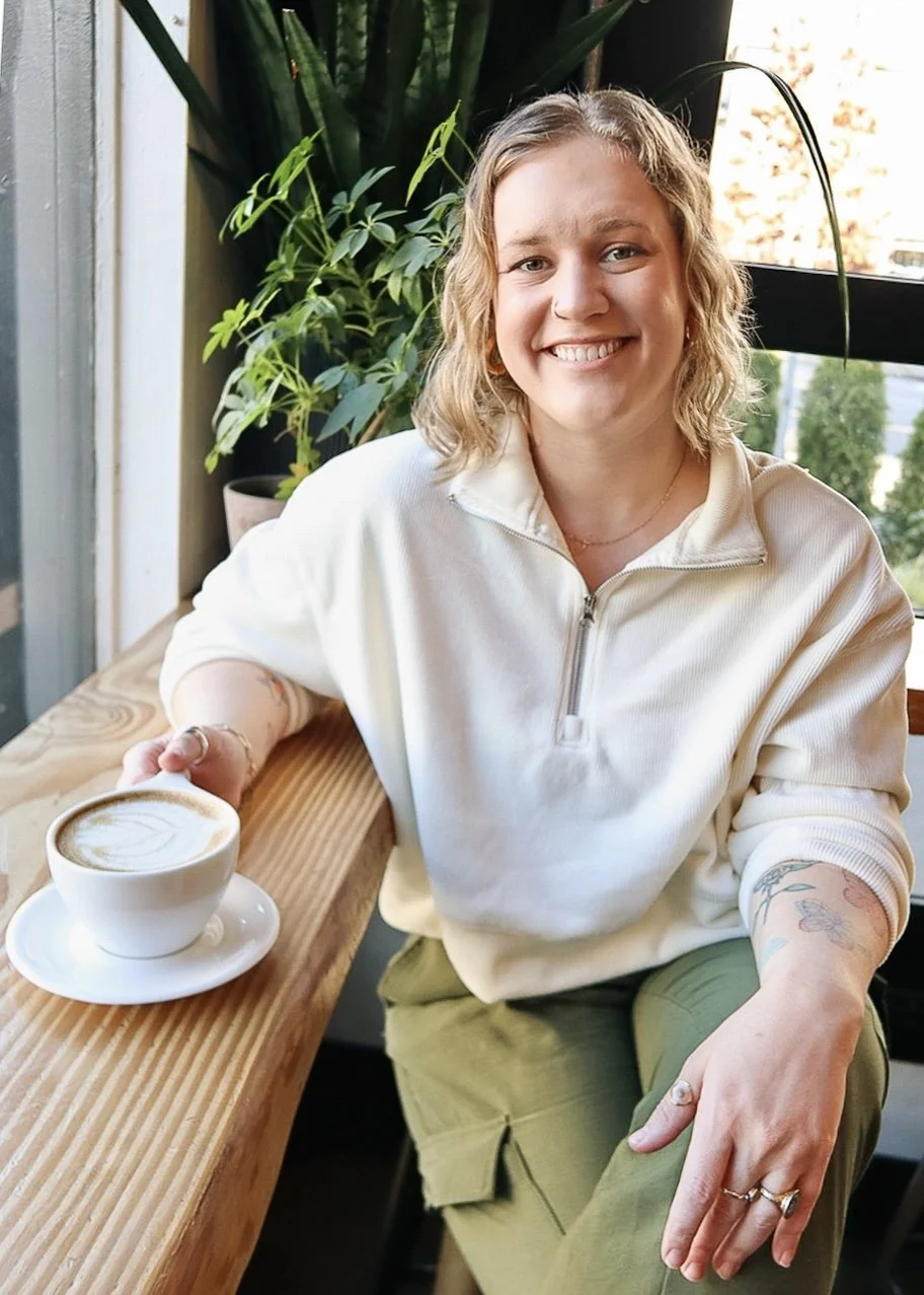 A woman sitting at a wooden table in a cafe, smiling and holding a cup of coffee with latte art. She has curly blonde hair, is wearing a cream-colored quarter-zip sweater, and has a tattoo on her left forearm. There are green plants and a window in the background.