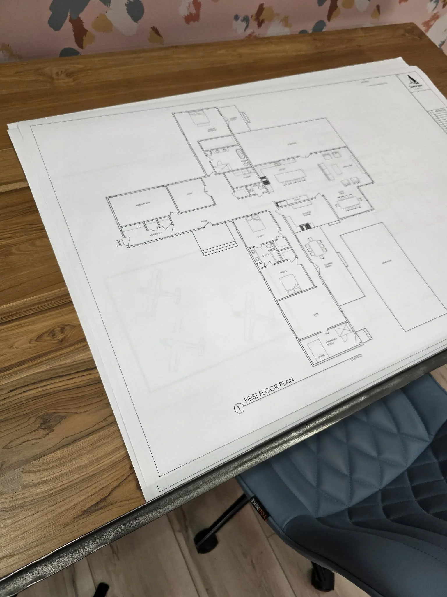 Architectural floor plan for a house on a large sheet of paper, placed on a wooden table with a chair nearby.