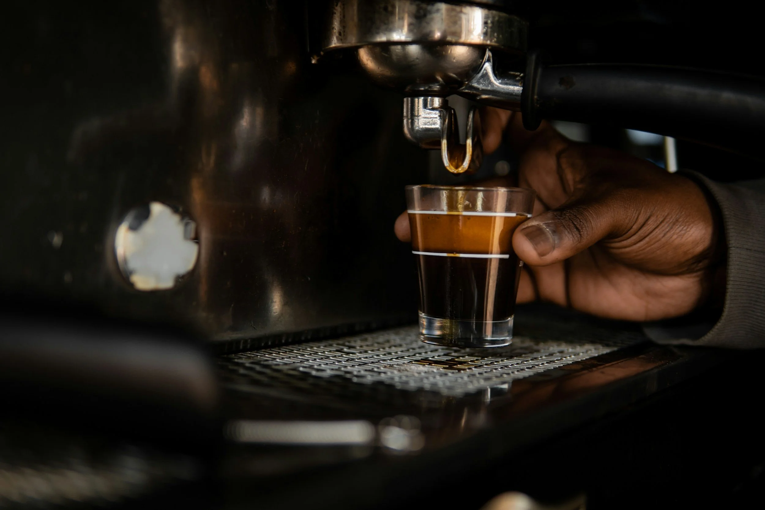 A person of color is holding an espresso shot underneath an espresso machine. The machine is actively pouring the drink into the glass.