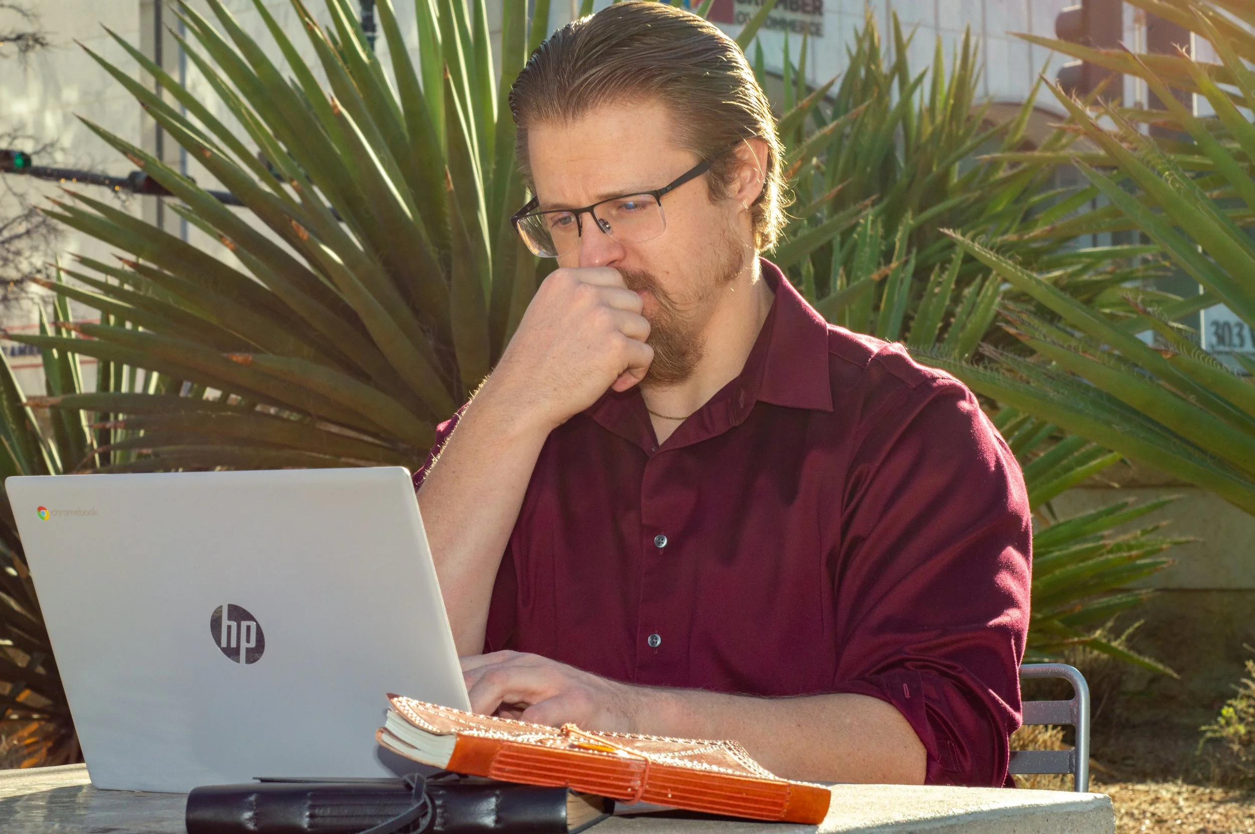 Business owner in a burgundy shirt using an HP laptop outdoors, with a thoughtful expression, sitting at a table with notebooks.