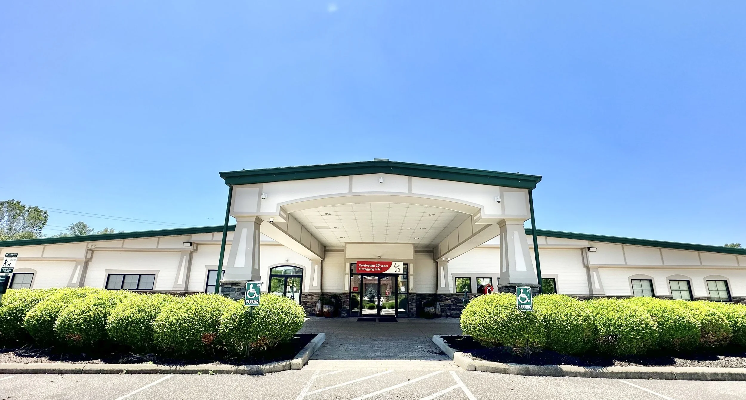 Front view Red Dog Pet Resort and Spa, developed by Circle Development, a commercial building with a covered entrance, surrounded by neatly trimmed bushes and parking spots with accessible parking signs. Clear blue sky overhead.