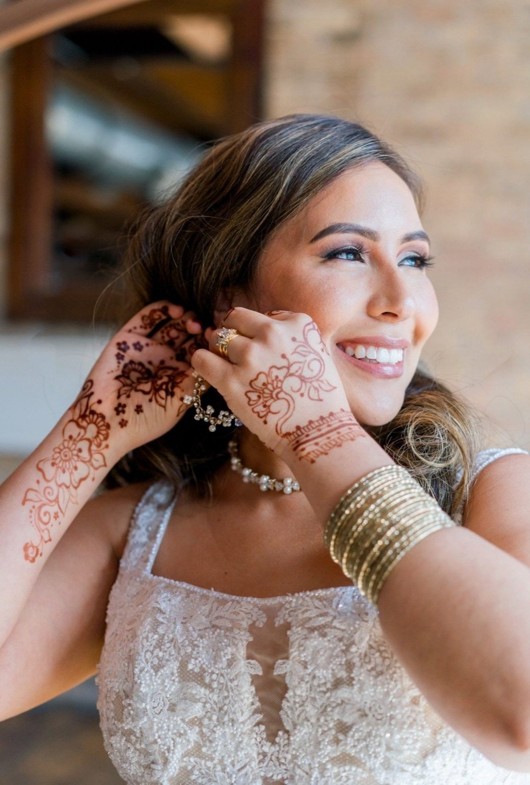 Smiling woman with henna decorated hands adjusting earrings, wearing a lace dress and gold bracelets.