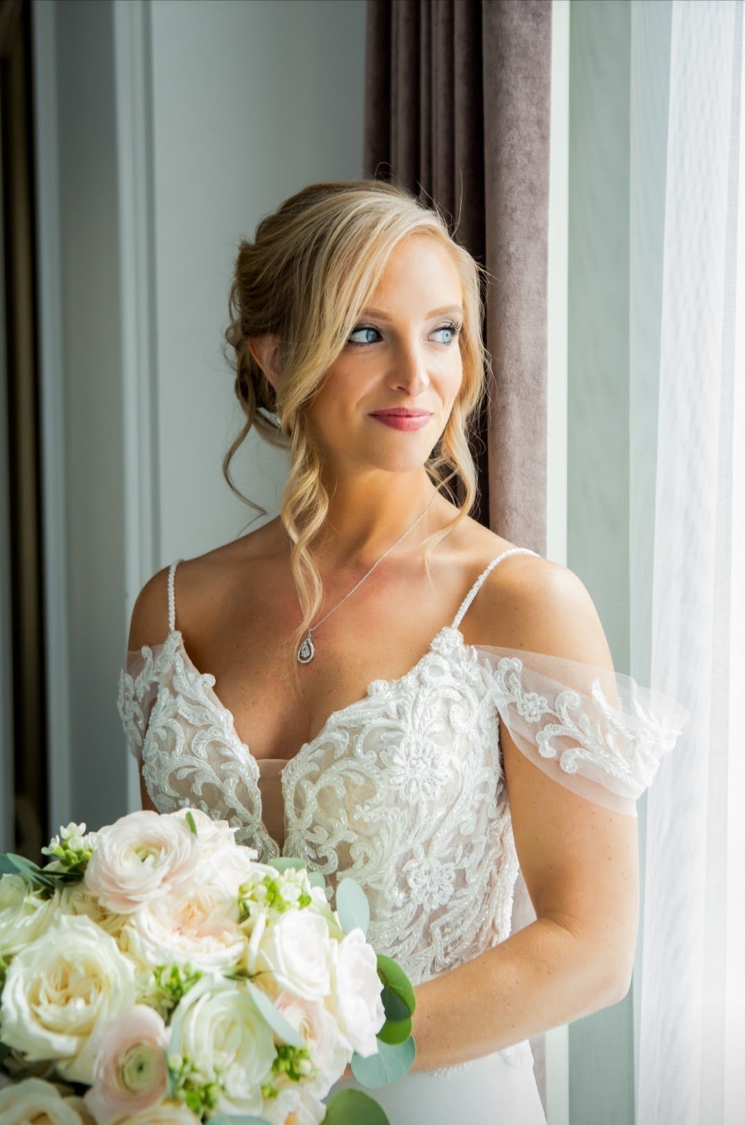 Bride in lace wedding dress holding a bouquet of white and blush roses, looking out the window.