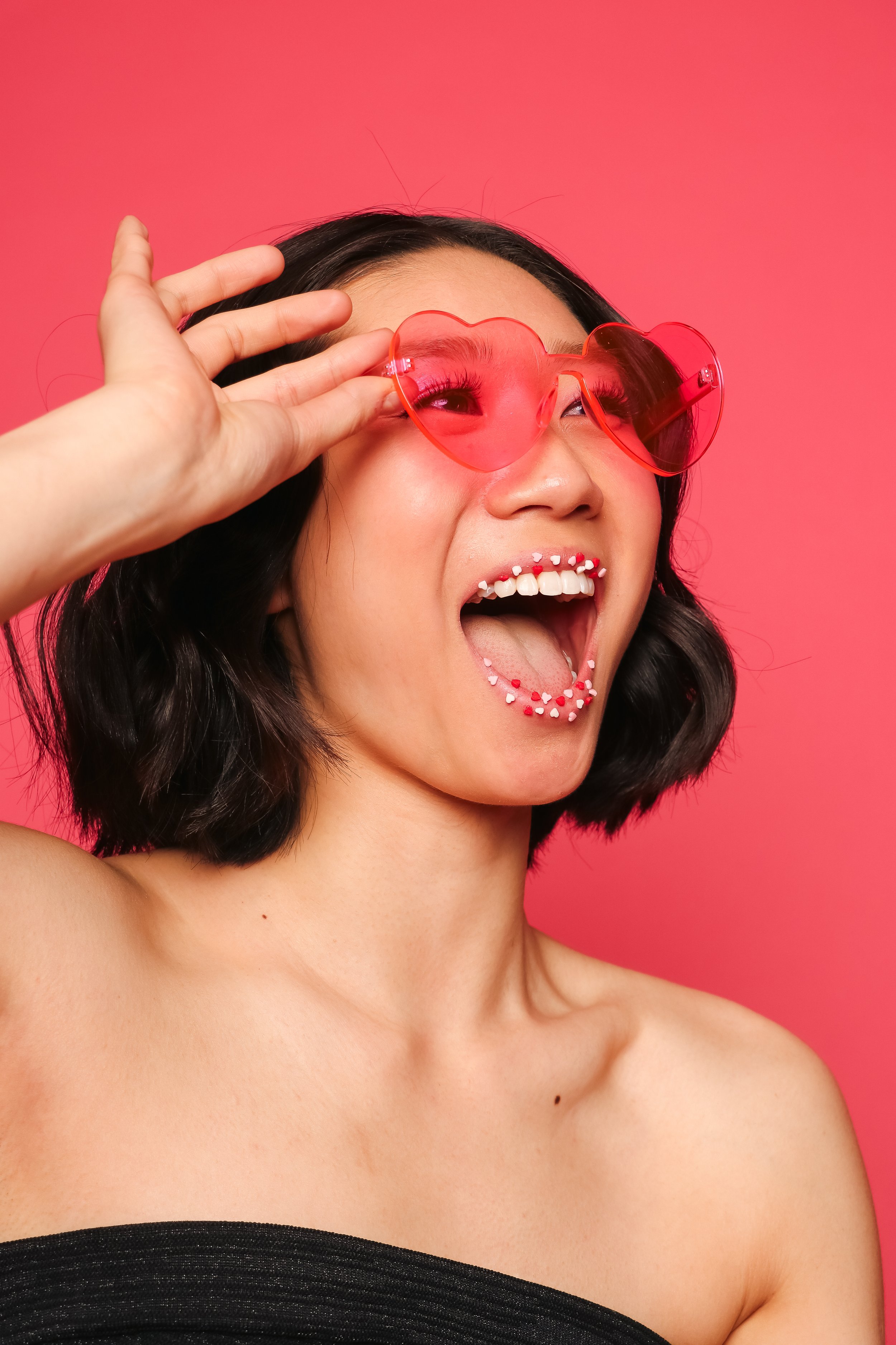 black haired woman wearing heart shaped glasses and black tube top against pink wall
