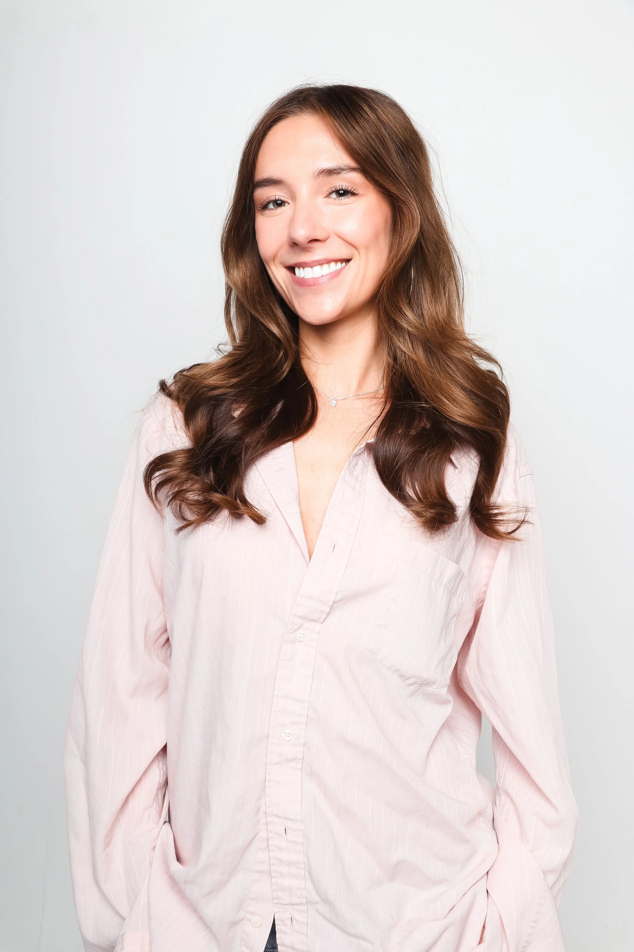brown haired woman with light pink button up shirt standing against white wall